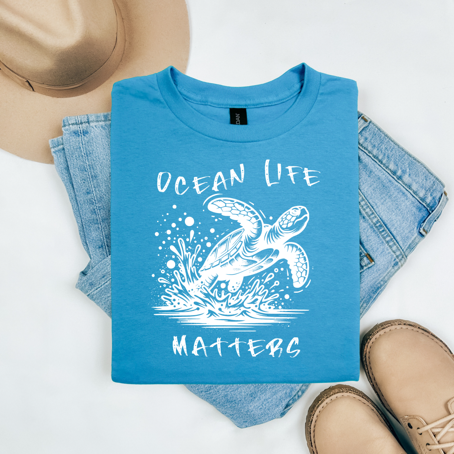 Gildan 5000 "Save The Ocean and The Sea Life In It" t shirt with a sea turtle splashing in water in a one color graphic. Picture on a flay lay mockup with pair of blue jeans, brown shoes and a hat accessory.