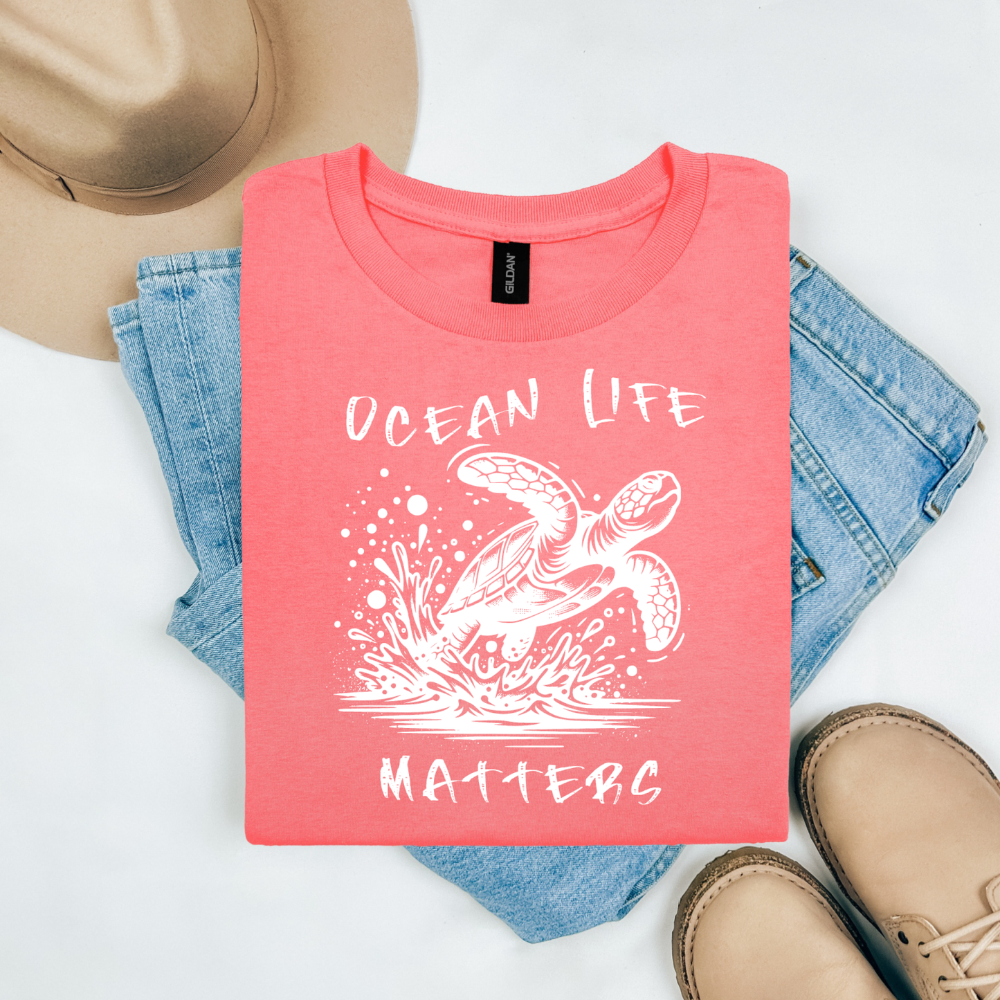 Gildan 5000 "Save The Ocean and The Sea Life In It" t shirt with a sea turtle splashing in water in a one color graphic. Picture on a flay lay mockup with pair of blue jeans, brown shoes and a hat accessory.