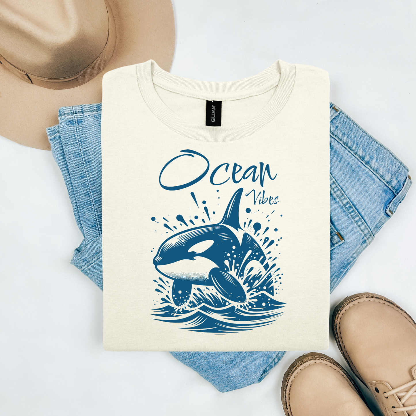 Gildan 5000 "Ocean Vibes" t shirt with a orca whale jumping out of water graphics in a one color graphic. Picture on a flay lay mockup with pair of blue jeans, brown shoes and a hat accessory.
