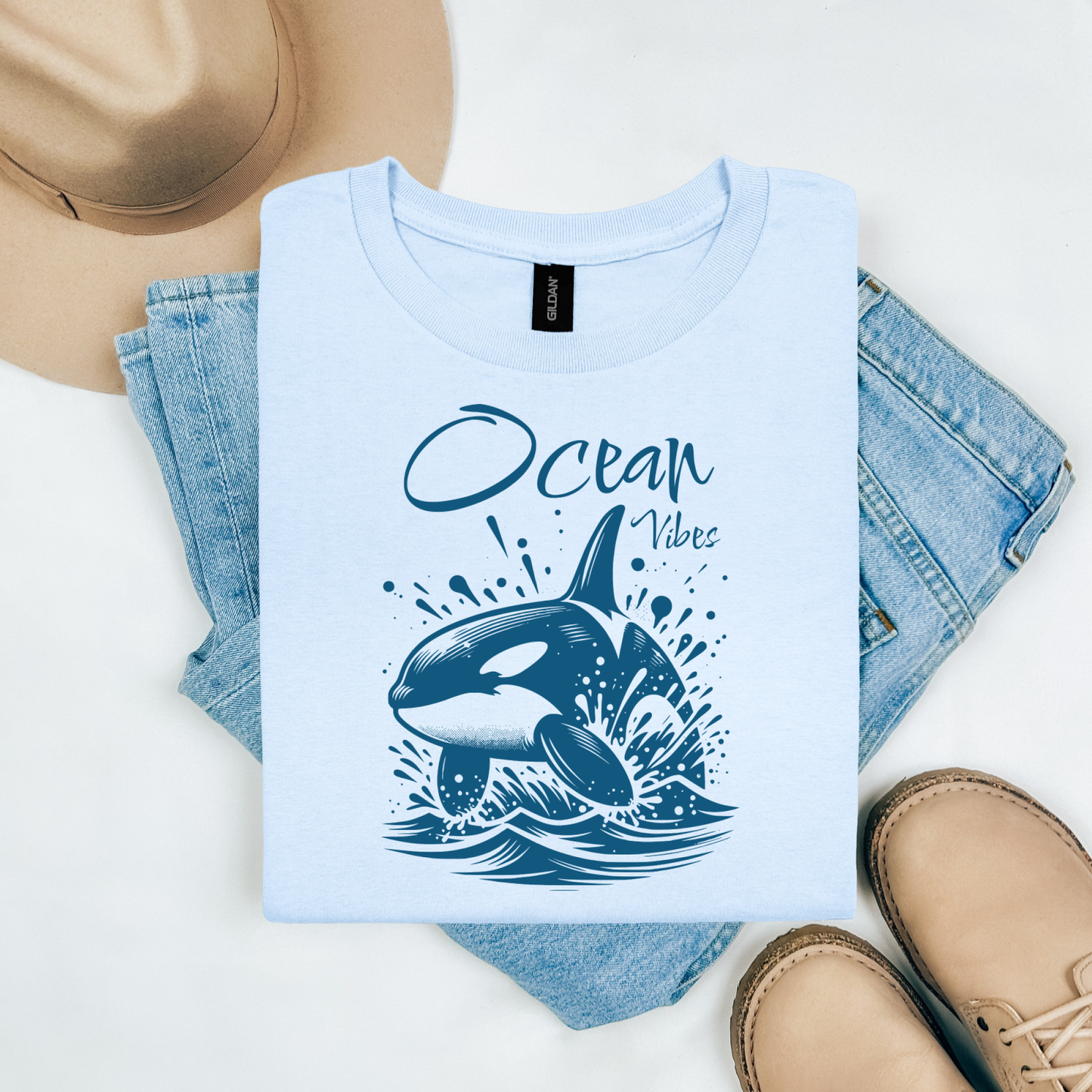Gildan 5000 "Ocean Vibes" t shirt with a orca whale jumping out of water graphics in a one color graphic. Picture on a flay lay mockup with pair of blue jeans, brown shoes and a hat accessory.