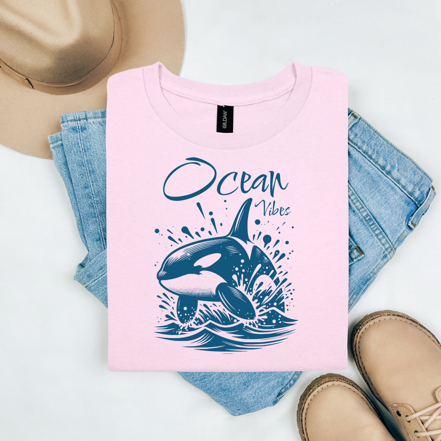 Gildan 5000 "Ocean Vibes" t shirt with a orca whale jumping out of water graphics in a one color graphic. Picture on a flay lay mockup with pair of blue jeans, brown shoes and a hat accessory.