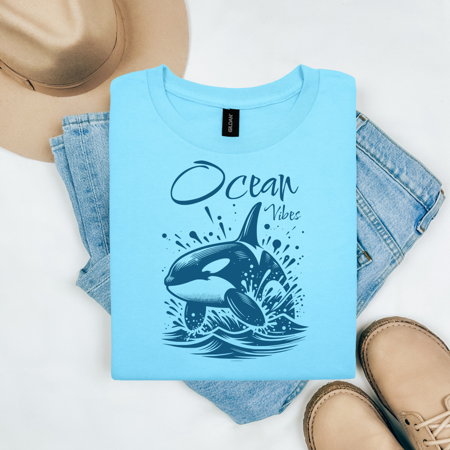 Gildan 5000 "Ocean Vibes" t shirt with a orca whale jumping out of water graphics in a one color graphic. Picture on a flay lay mockup with pair of blue jeans, brown shoes and a hat accessory.