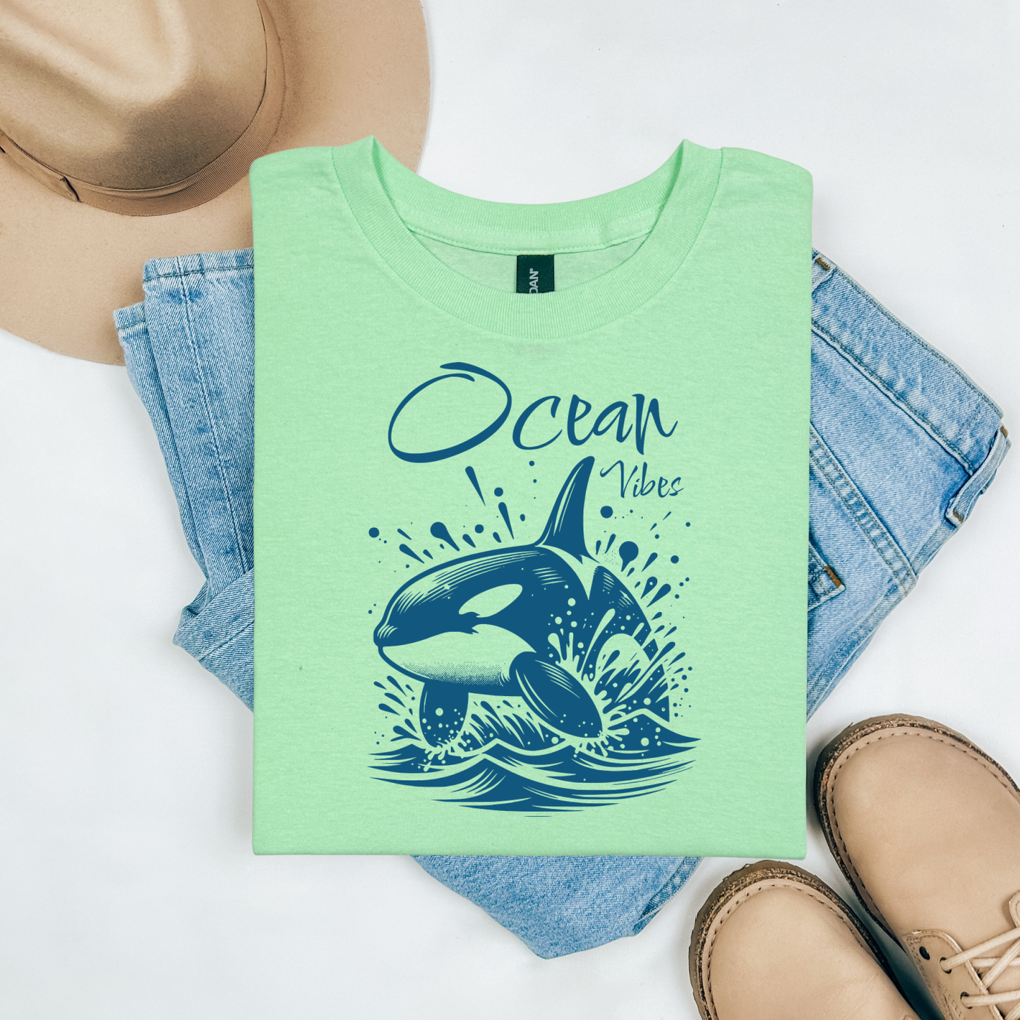 Gildan 5000 "Ocean Vibes" t shirt with a orca whale jumping out of water graphics in a one color graphic. Picture on a flay lay mockup with pair of blue jeans, brown shoes and a hat accessory.