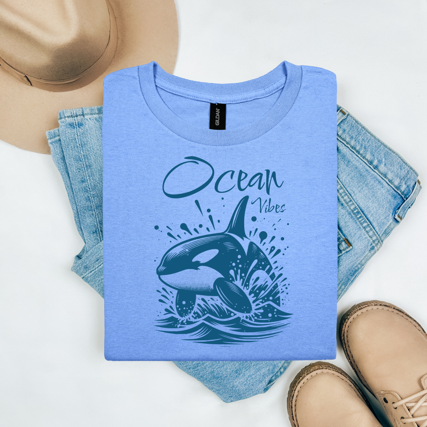 Gildan 5000 "Ocean Vibes" t shirt with a orca whale jumping out of water graphics in a one color graphic. Picture on a flay lay mockup with pair of blue jeans, brown shoes and a hat accessory.