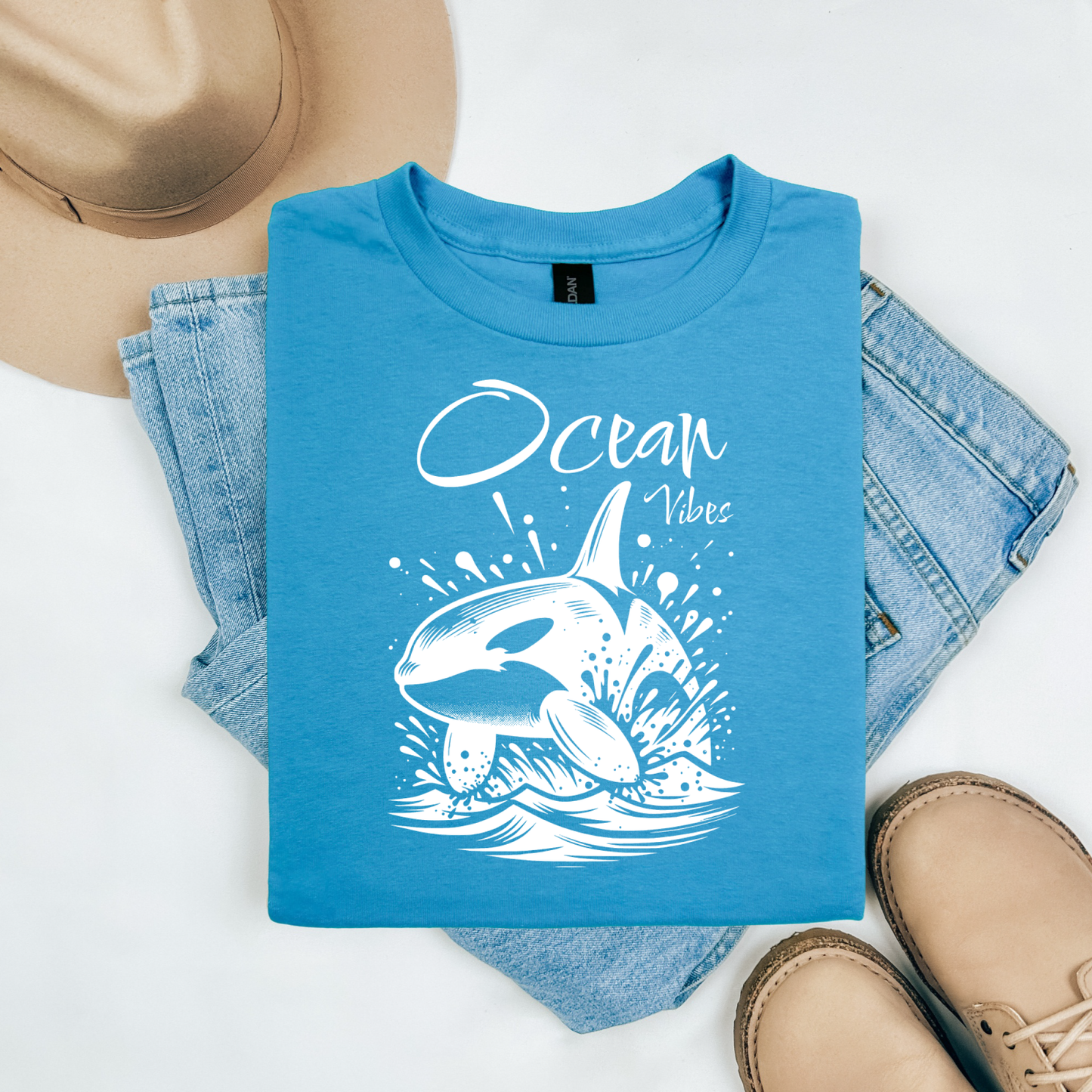 Gildan 5000 "Ocean Vibes" t shirt with a orca whale jumping out of water graphics in a one color graphic. Picture on a flay lay mockup with pair of blue jeans, brown shoes and a hat accessory.