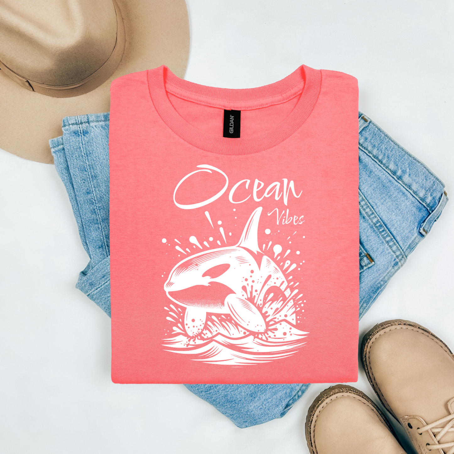 Gildan 5000 "Ocean Vibes" t shirt with a orca whale jumping out of water graphics in a one color graphic. Picture on a flay lay mockup with pair of blue jeans, brown shoes and a hat accessory.