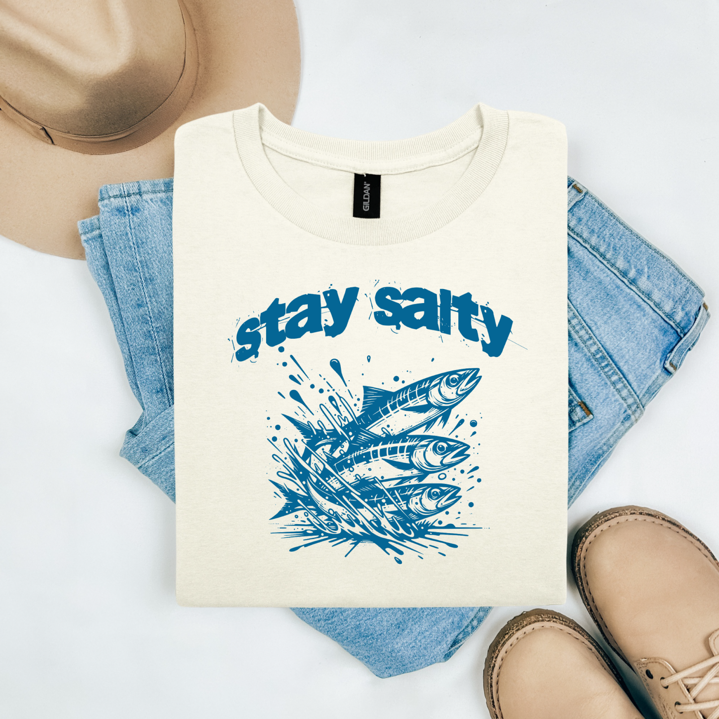 Gildan 5000 "Stay Salty" t shirt with Three Sardines jumping out of water in one color graphics. Picture on a flay lay mockup with pair of blue jeans, brown shoes and a hat accessory.