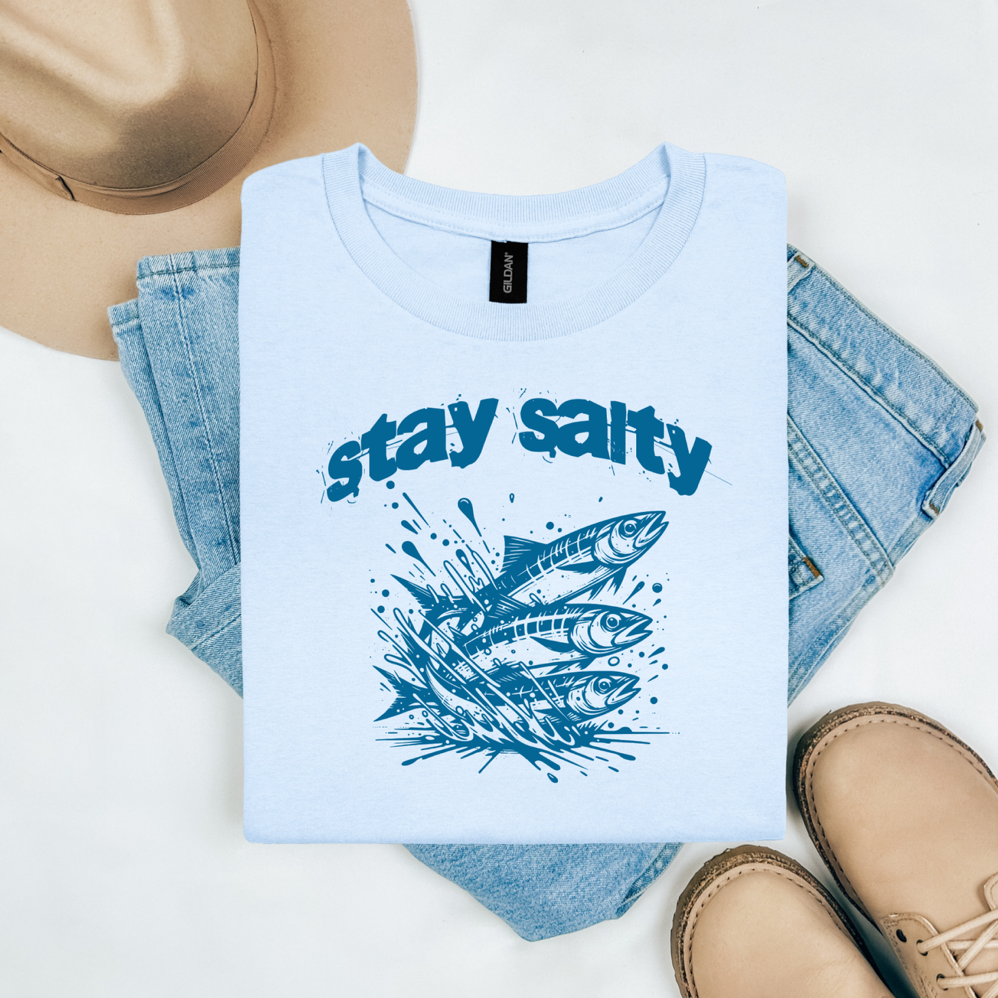 Gildan 5000 "Stay Salty" t shirt with Three Sardines jumping out of water in one color graphics. Picture on a flay lay mockup with pair of blue jeans, brown shoes and a hat accessory.
