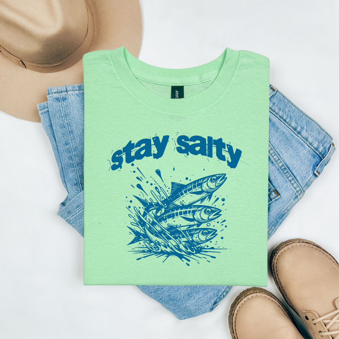 Gildan 5000 "Stay Salty" t shirt with Three Sardines jumping out of water in one color graphics. Picture on a flay lay mockup with pair of blue jeans, brown shoes and a hat accessory.