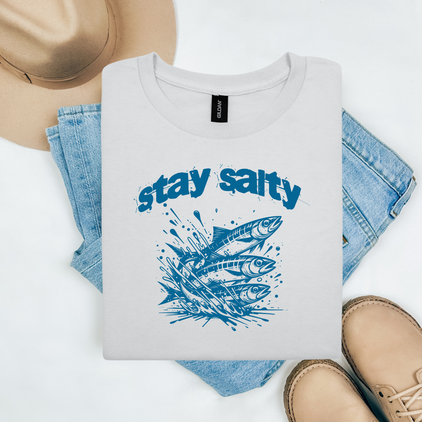 Gildan 5000 "Stay Salty" t shirt with Three Sardines jumping out of water in one color graphics. Picture on a flay lay mockup with pair of blue jeans, brown shoes and a hat accessory.