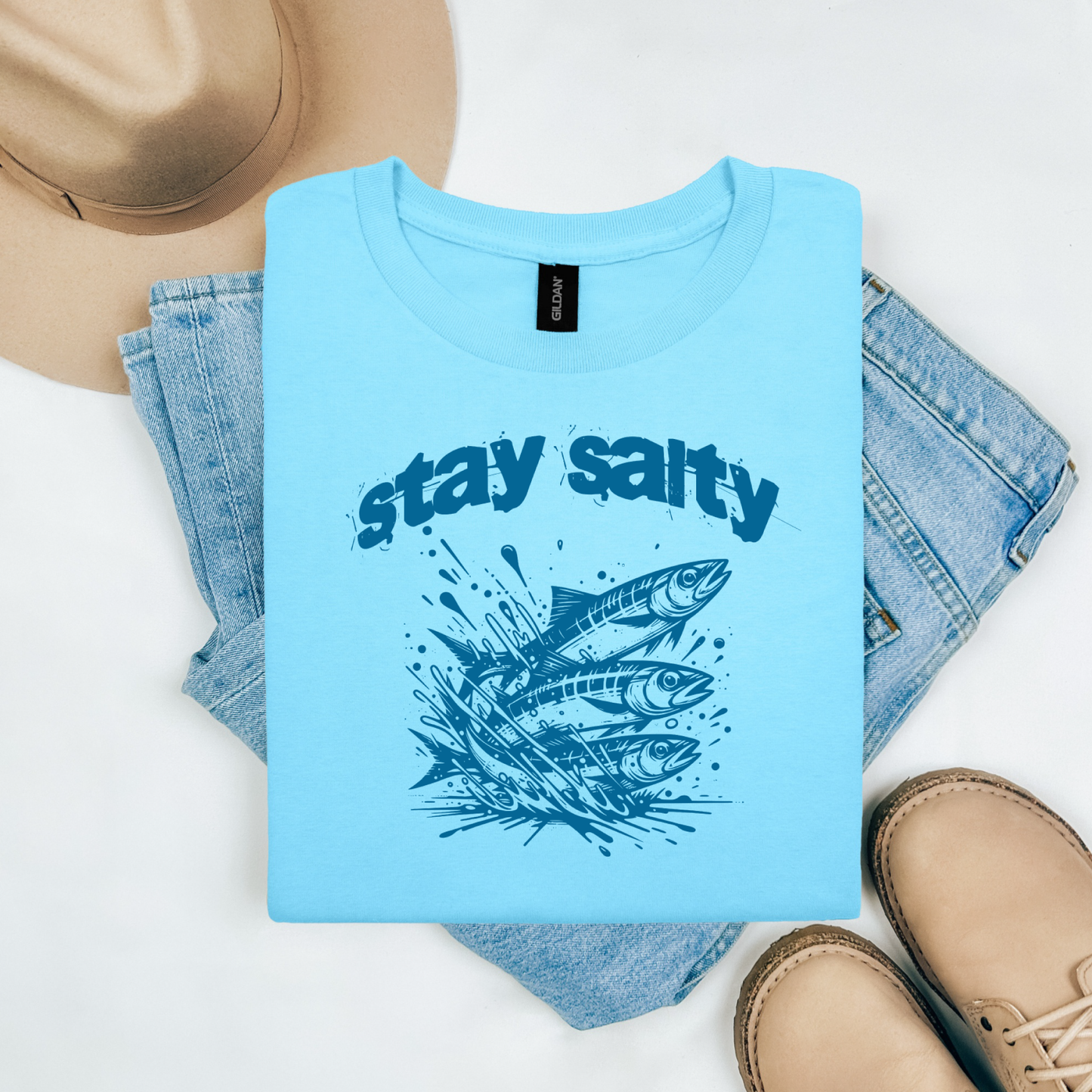 Gildan 5000 "Stay Salty" t shirt with Three Sardines jumping out of water in one color graphics. Picture on a flay lay mockup with pair of blue jeans, brown shoes and a hat accessory.