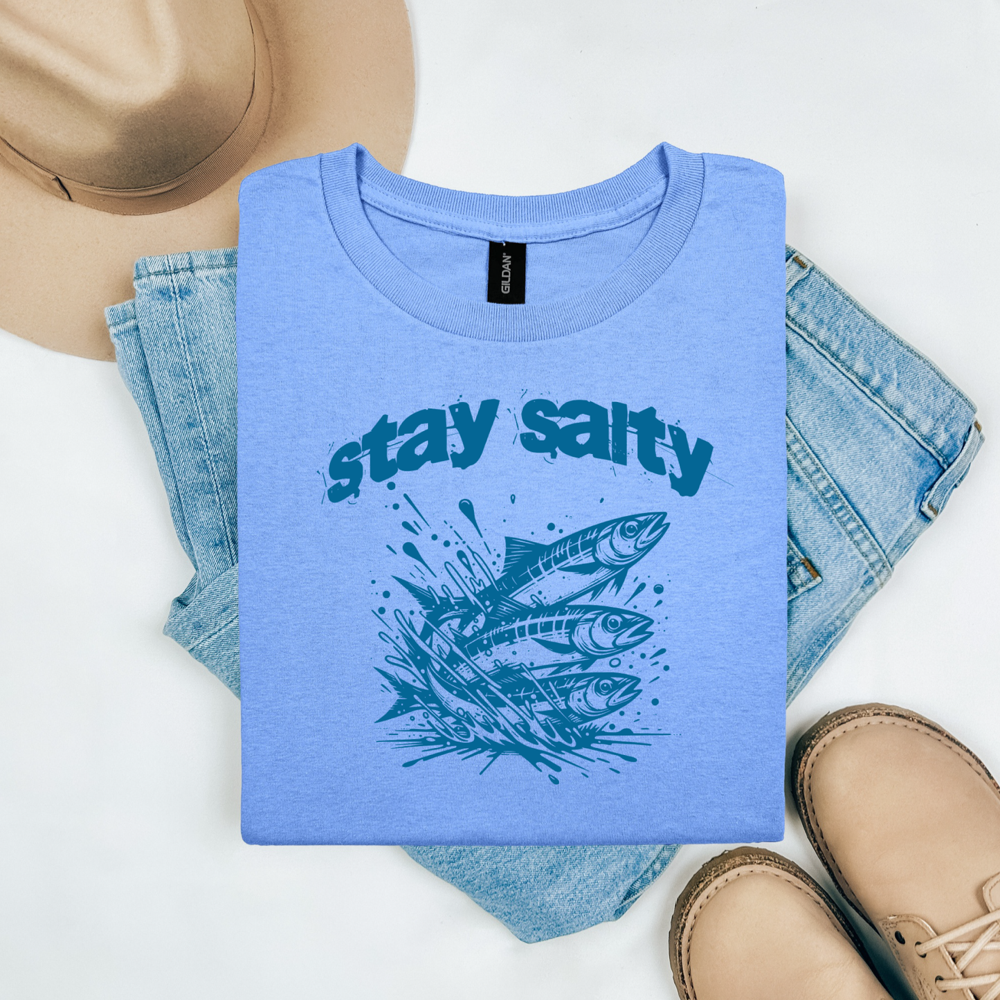 Gildan 5000 "Stay Salty" t shirt with Three Sardines jumping out of water in one color graphics. Picture on a flay lay mockup with pair of blue jeans, brown shoes and a hat accessory.