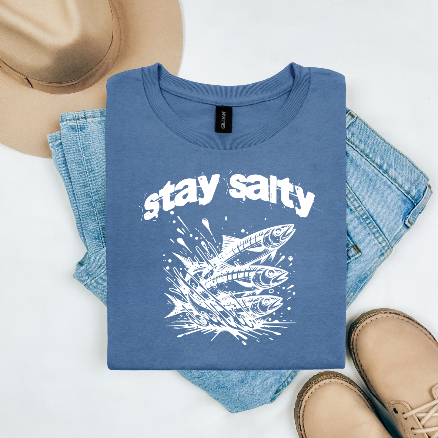 Gildan 5000 "Stay Salty" t shirt with Three Sardines jumping out of water in one color graphics. Picture on a flay lay mockup with pair of blue jeans, brown shoes and a hat accessory.