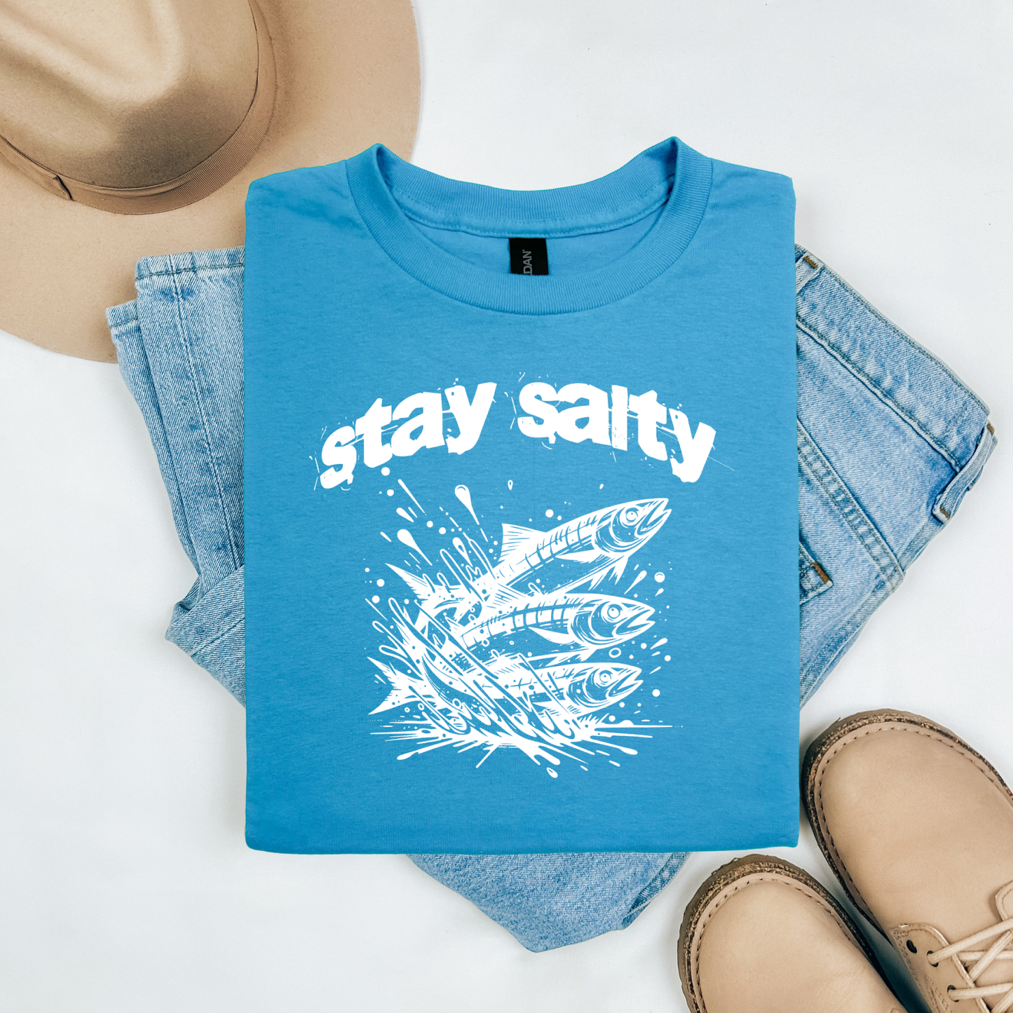 Gildan 5000 "Stay Salty" t shirt with Three Sardines jumping out of water in one color graphics. Picture on a flay lay mockup with pair of blue jeans, brown shoes and a hat accessory.