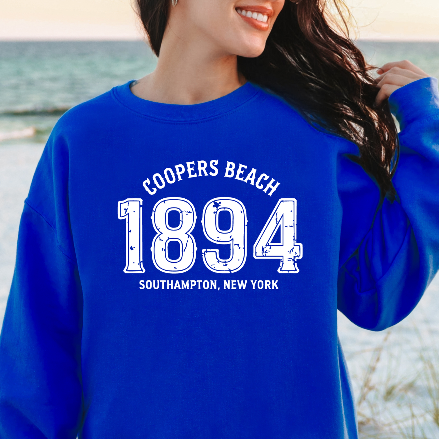 Person wearing a blue sweatshirt with 'Coopers Beach 1894' text, standing by a body of water.