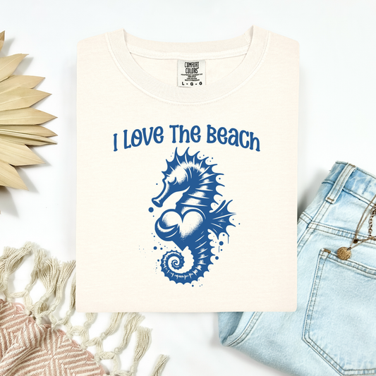 Comfort Colors 1717 "I Love The Beach" t shirt with a seahorse holding a heart graphics in one color. Picture on a flay lay mockup with pair of blue jeans and blanket and a wicker sun shaped accessory.