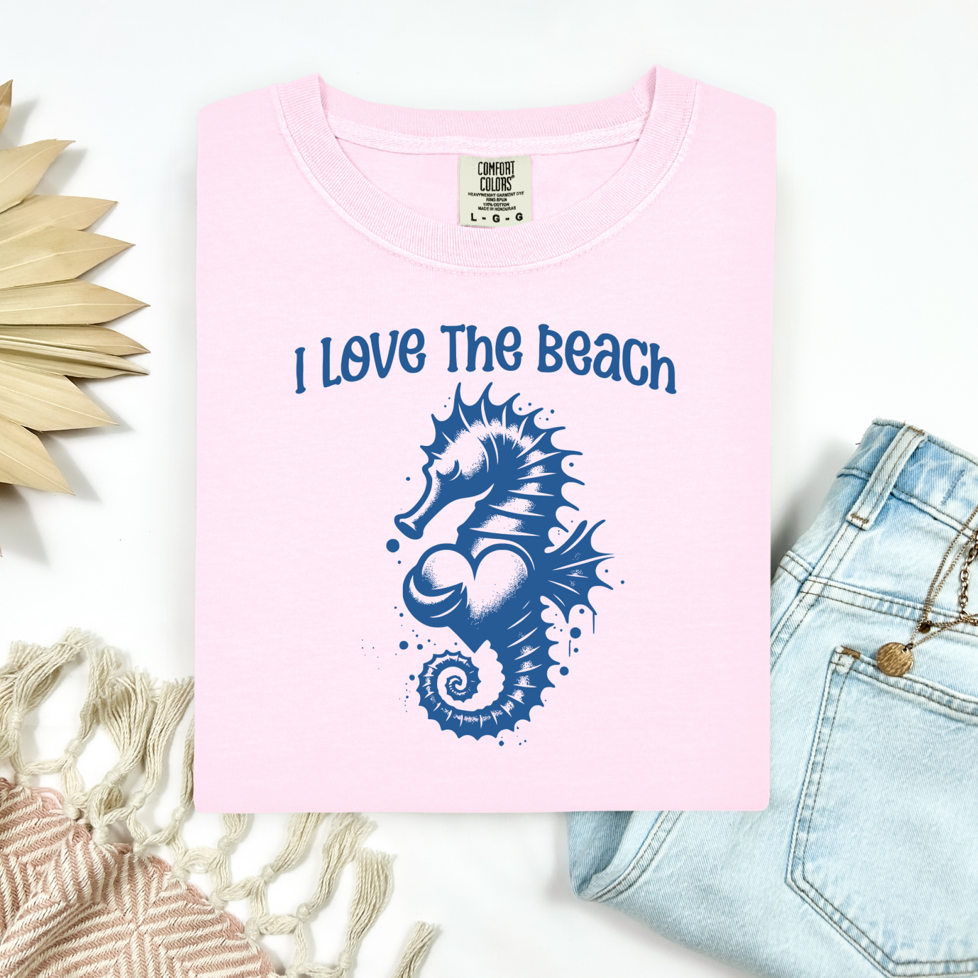 Comfort Colors 1717 "I Love The Beach" t shirt with a seahorse holding a heart graphics in one color. Picture on a flay lay mockup with pair of blue jeans and blanket and a wicker sun shaped accessory.