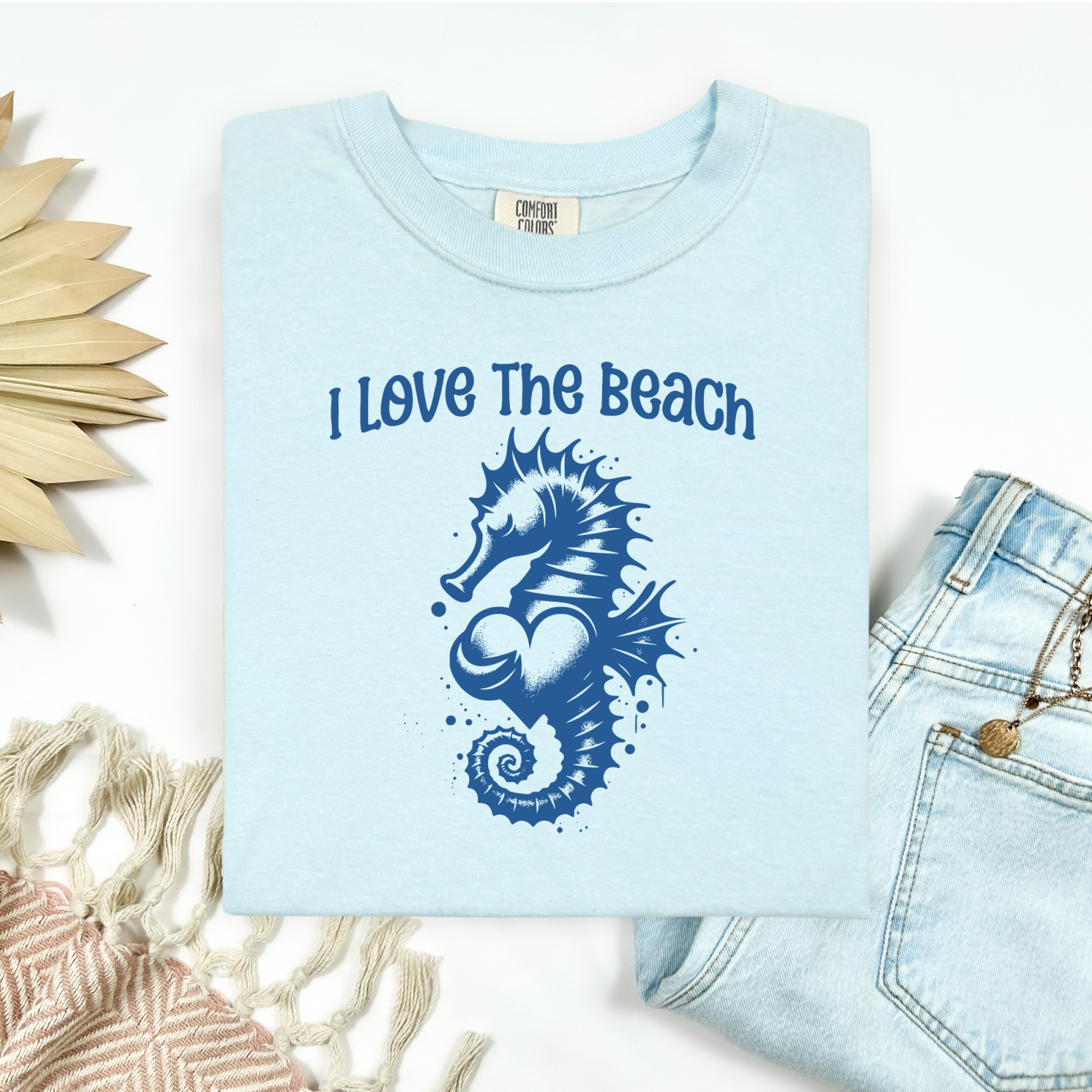 Comfort Colors 1717 "I Love The Beach" t shirt with a seahorse holding a heart graphics in one color. Picture on a flay lay mockup with pair of blue jeans and blanket and a wicker sun shaped accessory.