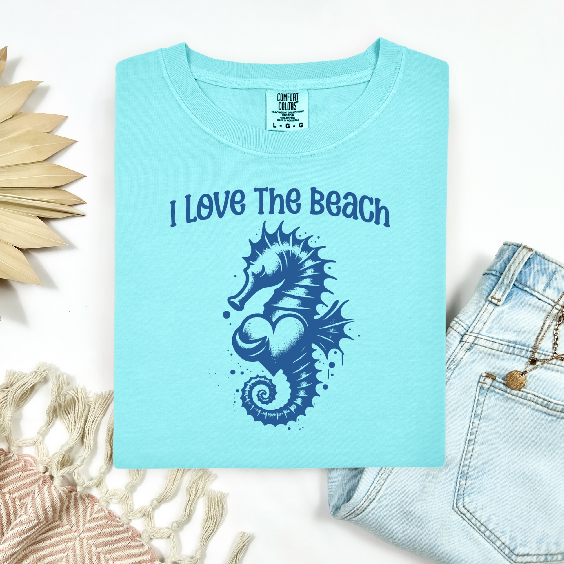 Comfort Colors 1717 "I Love The Beach" t shirt with a seahorse holding a heart graphics in one color. Picture on a flay lay mockup with pair of blue jeans and blanket and a wicker sun shaped accessory.