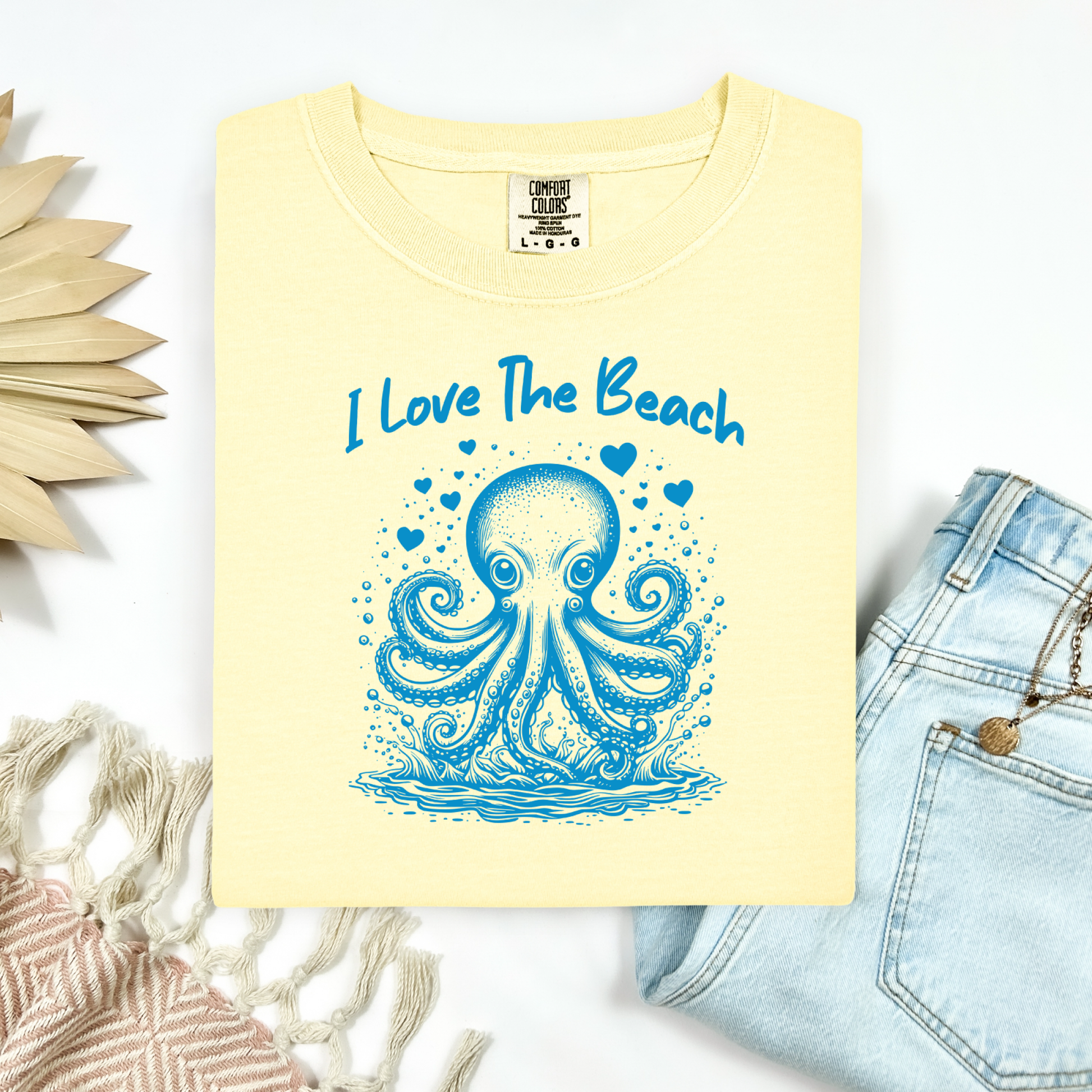 The Comfort Colors 1717 "I Love The Beach" T-shirt features a charming one-color graphic of an octopus surrounded by hearts, bringing a touch of whimsy and coastal charm. Displayed in a flat lay mockup, the tee is styled with a pair of blue jeans, a cozy blanket, and a wicker sun-shaped accessory—perfect for showcasing a relaxed, beachy vibe.