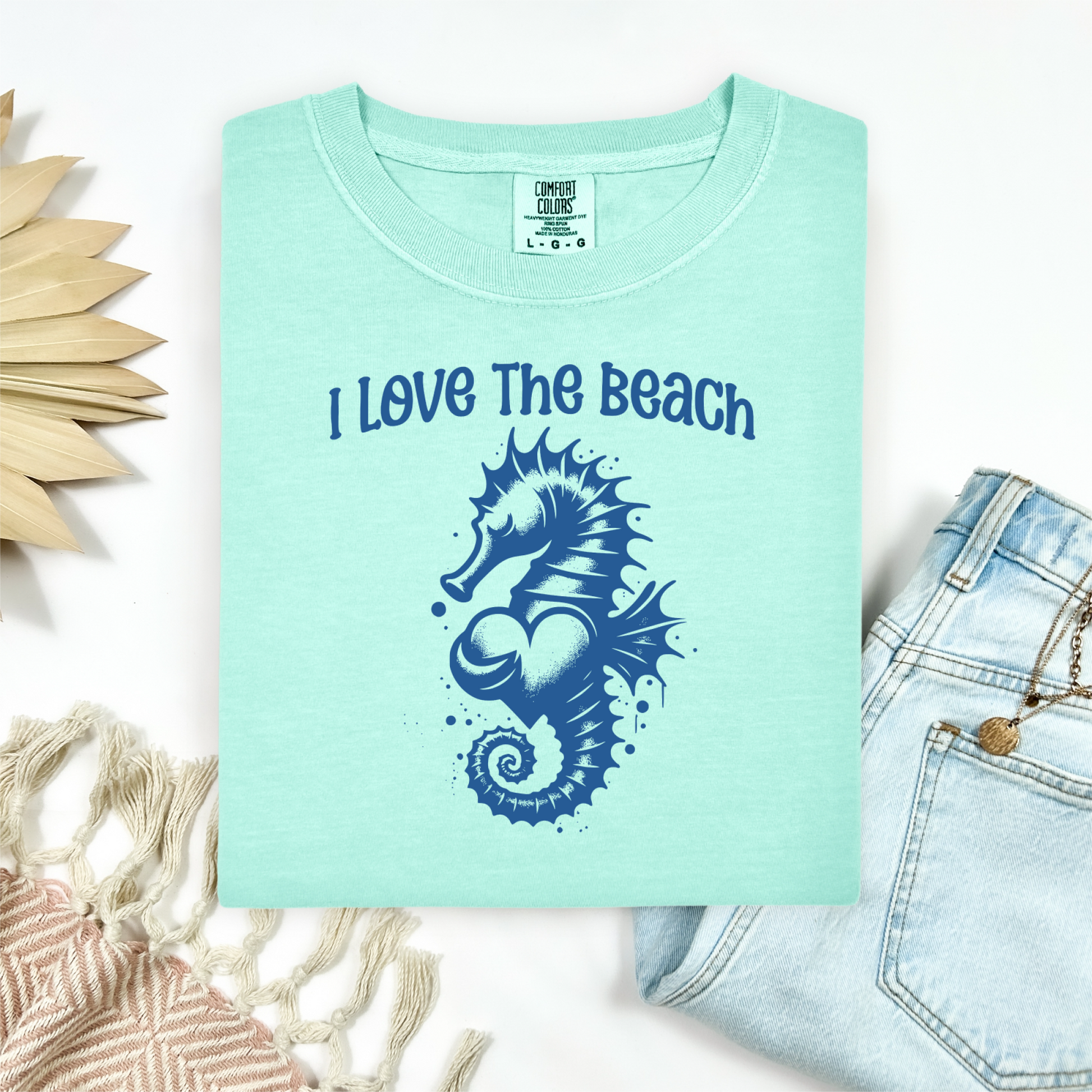 Comfort Colors 1717 "I Love The Beach" t shirt with a seahorse holding a heart graphics in one color. Picture on a flay lay mockup with pair of blue jeans and blanket and a wicker sun shaped accessory.