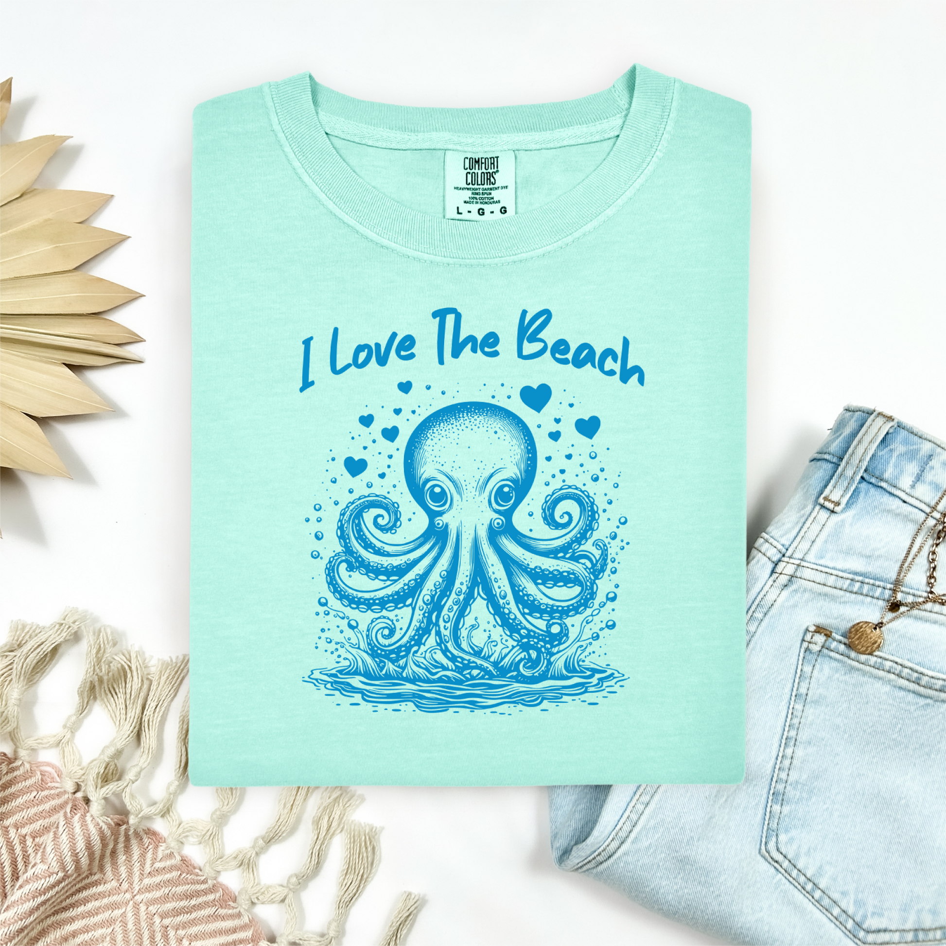 The Comfort Colors 1717 "I Love The Beach" T-shirt features a charming one-color graphic of an octopus surrounded by hearts, bringing a touch of whimsy and coastal charm. Displayed in a flat lay mockup, the tee is styled with a pair of blue jeans, a cozy blanket, and a wicker sun-shaped accessory—perfect for showcasing a relaxed, beachy vibe.