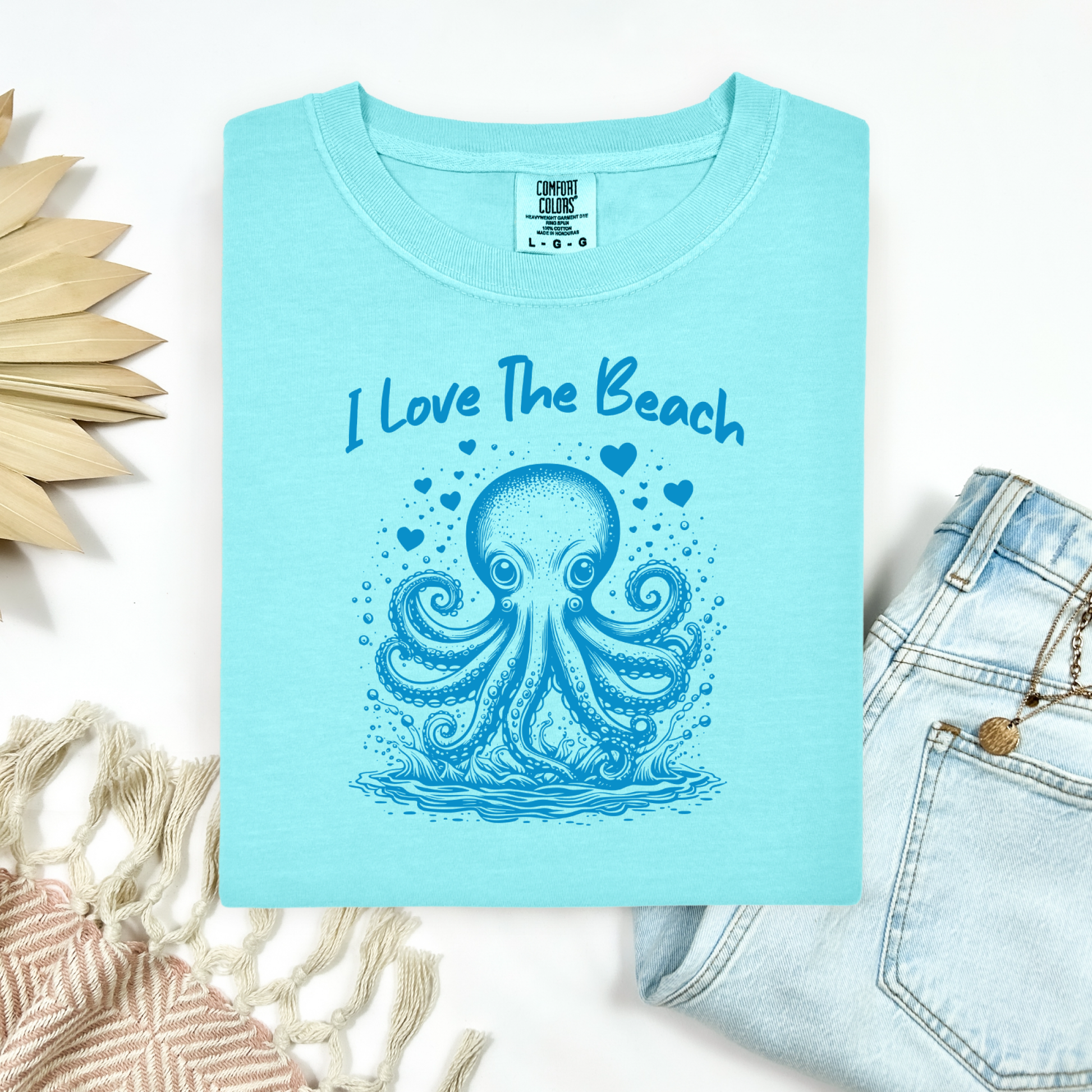 The Comfort Colors 1717 "I Love The Beach" T-shirt features a charming one-color graphic of an octopus surrounded by hearts, bringing a touch of whimsy and coastal charm. Displayed in a flat lay mockup, the tee is styled with a pair of blue jeans, a cozy blanket, and a wicker sun-shaped accessory—perfect for showcasing a relaxed, beachy vibe.