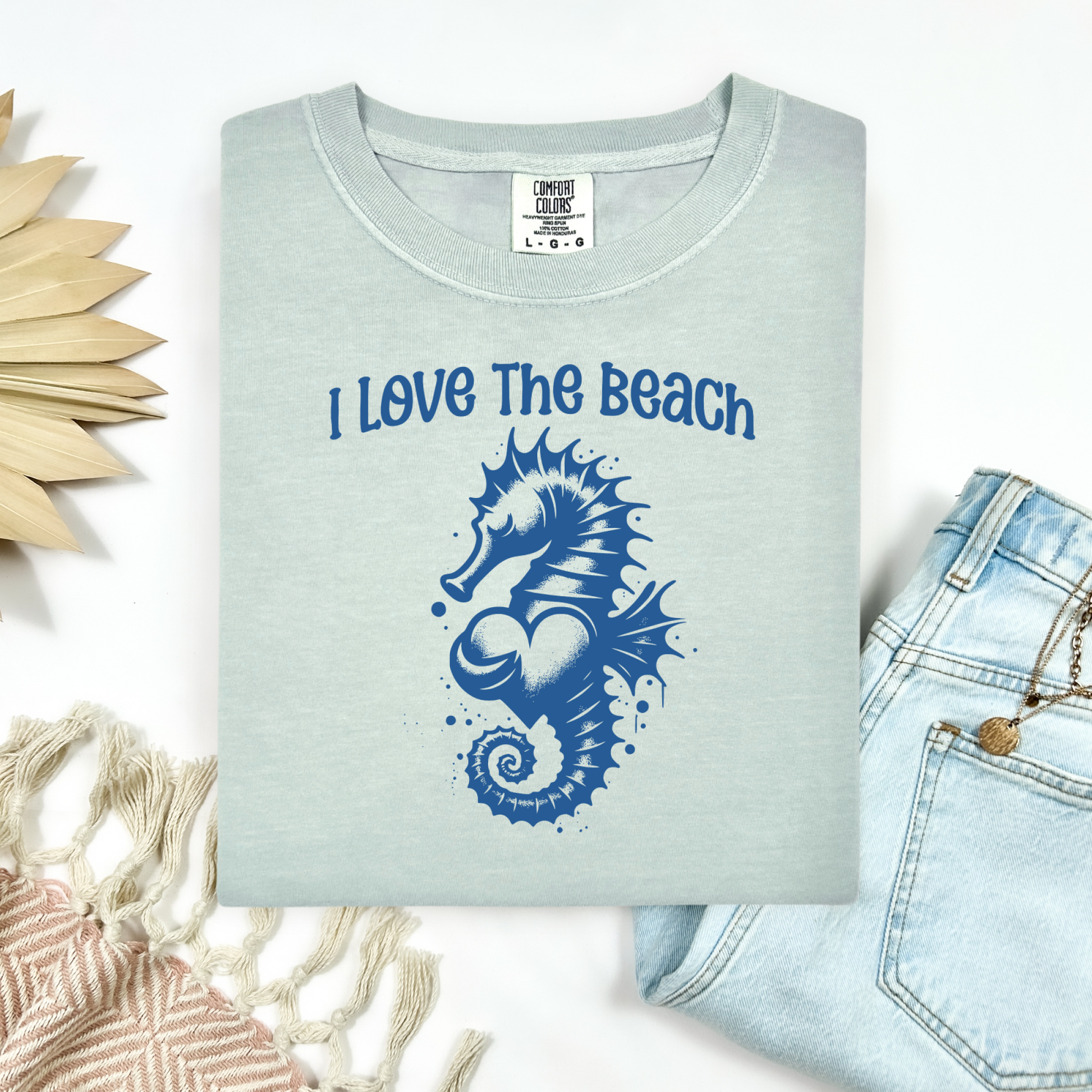 Comfort Colors 1717 "I Love The Beach" t shirt with a seahorse holding a heart graphics in one color. Picture on a flay lay mockup with pair of blue jeans and blanket and a wicker sun shaped accessory.