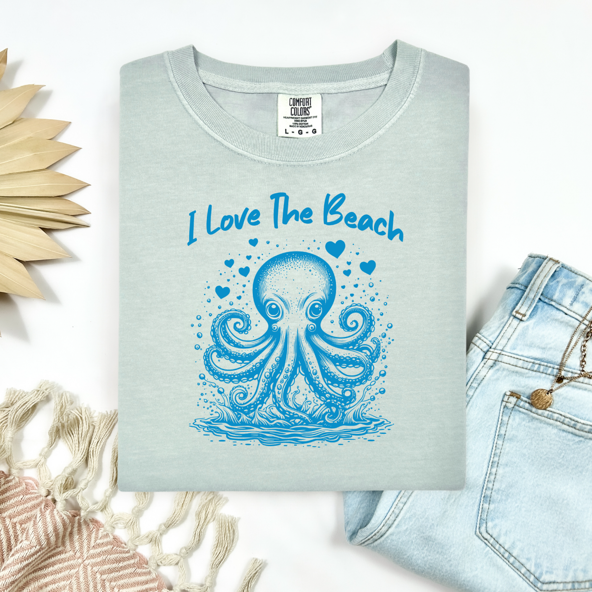 The Comfort Colors 1717 "I Love The Beach" T-shirt features a charming one-color graphic of an octopus surrounded by hearts, bringing a touch of whimsy and coastal charm. Displayed in a flat lay mockup, the tee is styled with a pair of blue jeans, a cozy blanket, and a wicker sun-shaped accessory—perfect for showcasing a relaxed, beachy vibe.