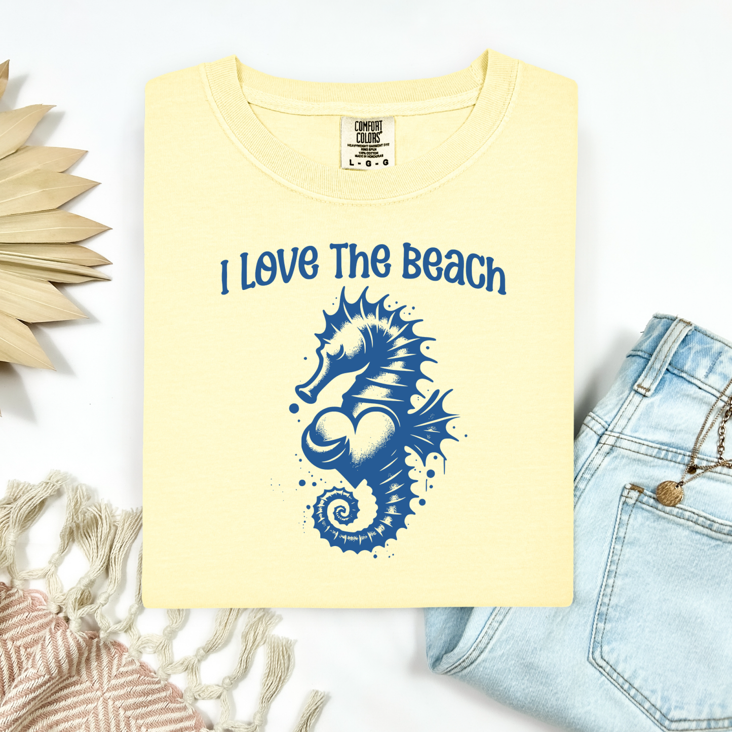 Comfort Colors 1717 "I Love The Beach" t shirt with a seahorse holding a heart graphics in one color. Picture on a flay lay mockup with pair of blue jeans and blanket and a wicker sun shaped accessory.