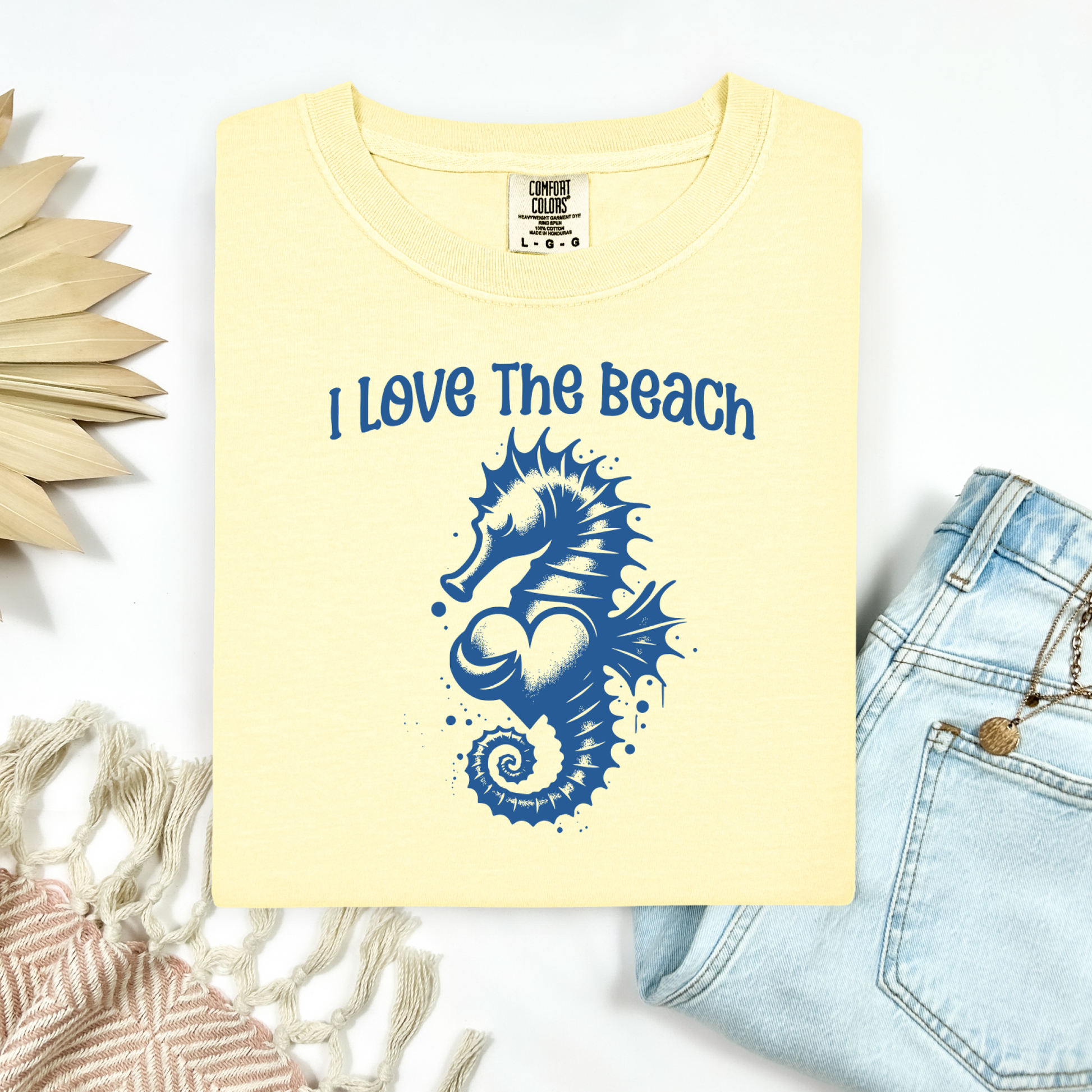 Comfort Colors 1717 "I Love The Beach" t shirt with a seahorse holding a heart graphics in one color. Picture on a flay lay mockup with pair of blue jeans and blanket and a wicker sun shaped accessory.