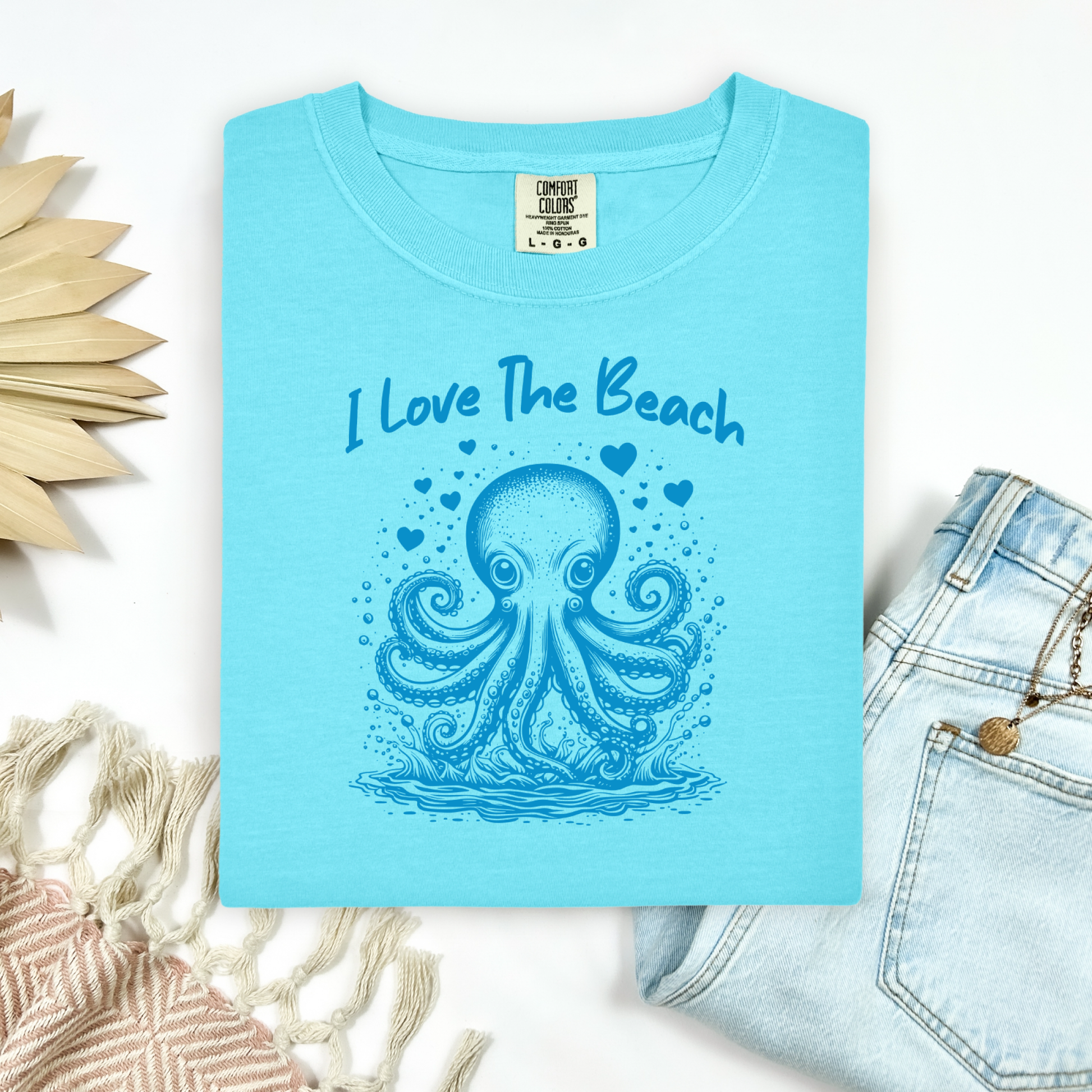 The Comfort Colors 1717 "I Love The Beach" T-shirt features a charming one-color graphic of an octopus surrounded by hearts, bringing a touch of whimsy and coastal charm. Displayed in a flat lay mockup, the tee is styled with a pair of blue jeans, a cozy blanket, and a wicker sun-shaped accessory—perfect for showcasing a relaxed, beachy vibe.