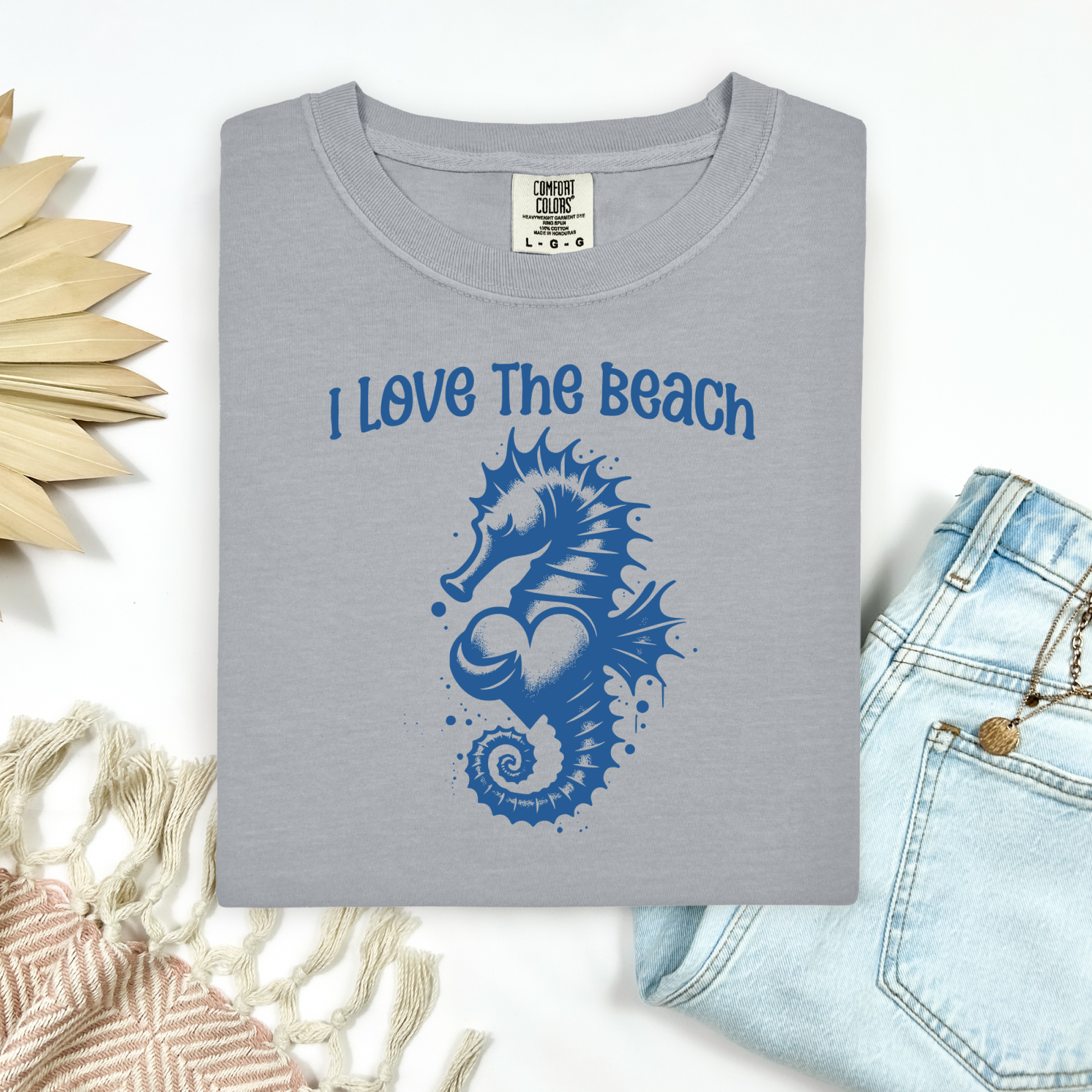 Comfort Colors 1717 "I Love The Beach" t shirt with a seahorse holding a heart graphics in one color. Picture on a flay lay mockup with pair of blue jeans and blanket and a wicker sun shaped accessory.