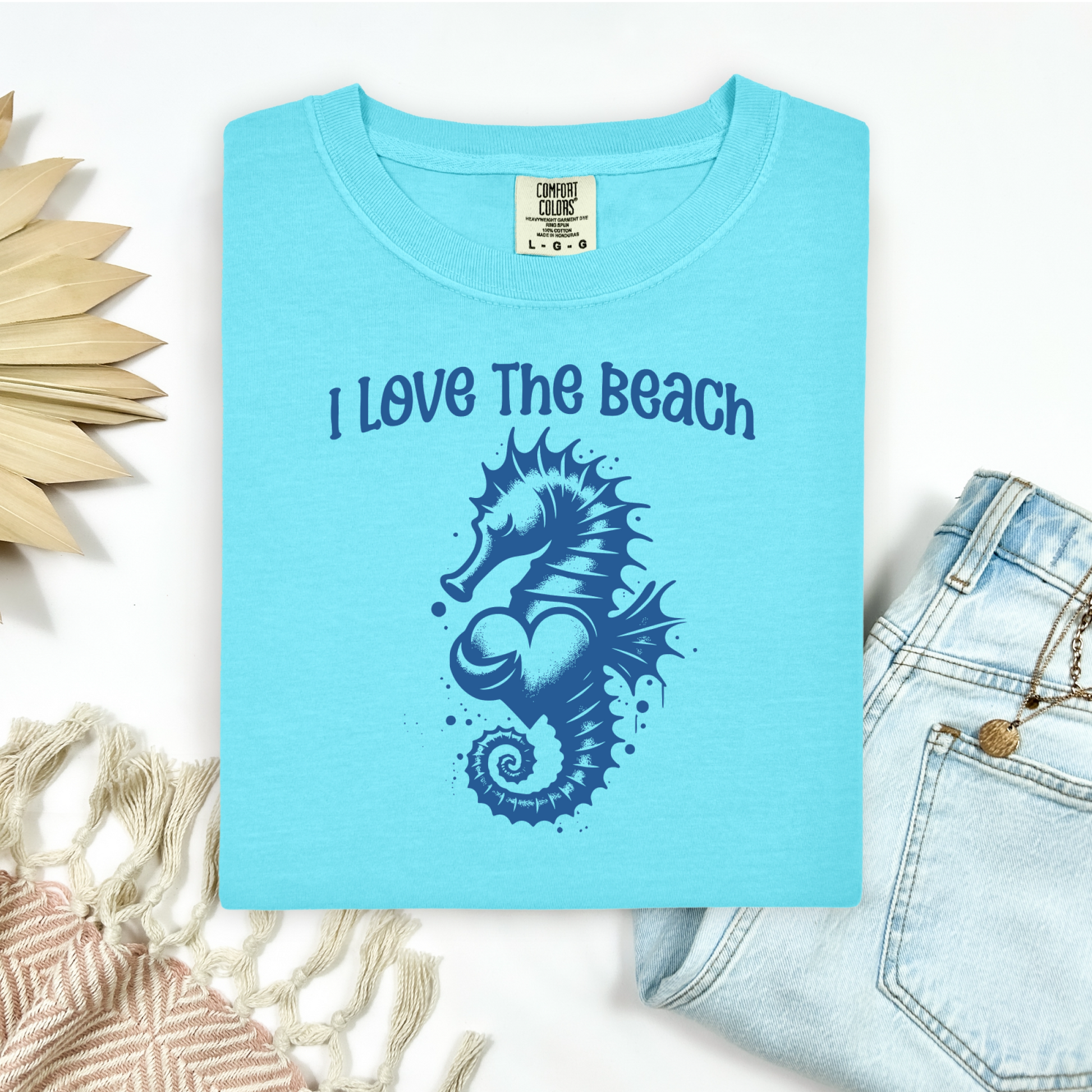 Comfort Colors 1717 "I Love The Beach" t shirt with a seahorse holding a heart graphics in one color. Picture on a flay lay mockup with pair of blue jeans and blanket and a wicker sun shaped accessory.