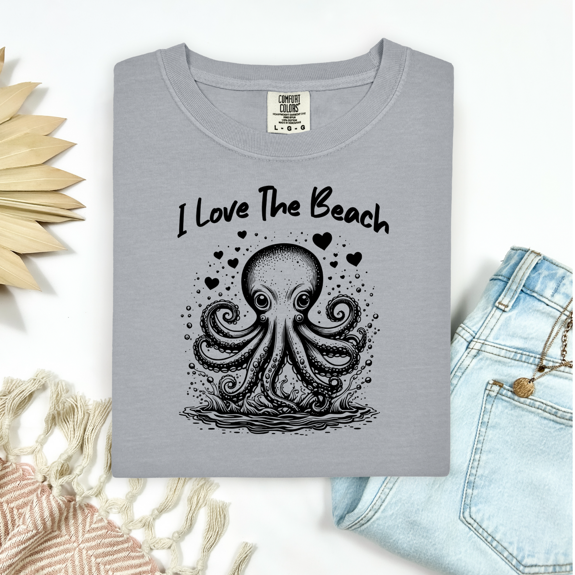 The Comfort Colors 1717 "I Love The Beach" T-shirt features a charming one-color graphic of an octopus surrounded by hearts, bringing a touch of whimsy and coastal charm. Displayed in a flat lay mockup, the tee is styled with a pair of blue jeans, a cozy blanket, and a wicker sun-shaped accessory—perfect for showcasing a relaxed, beachy vibe.