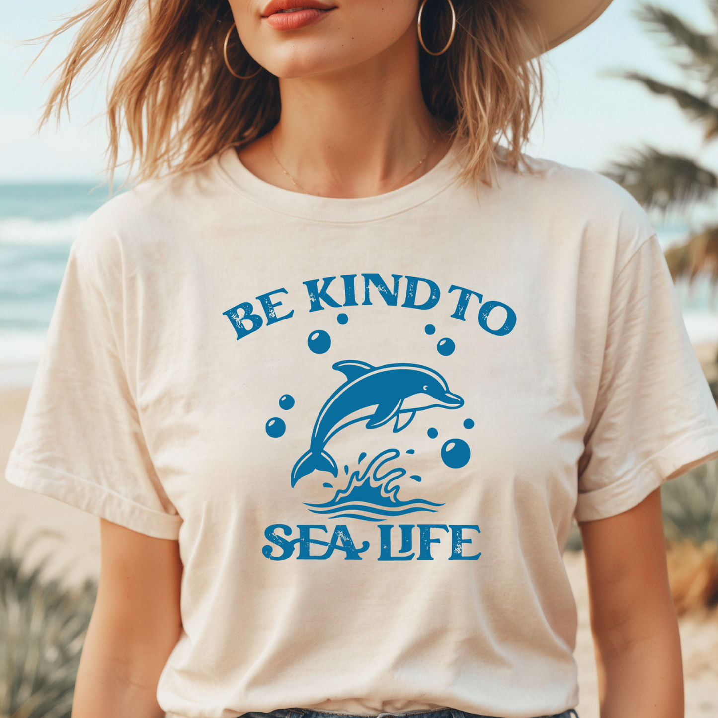 Person wearing a t-shirt with a dolphin graphic and 'Be Kind to Sea Life' text on a beach.