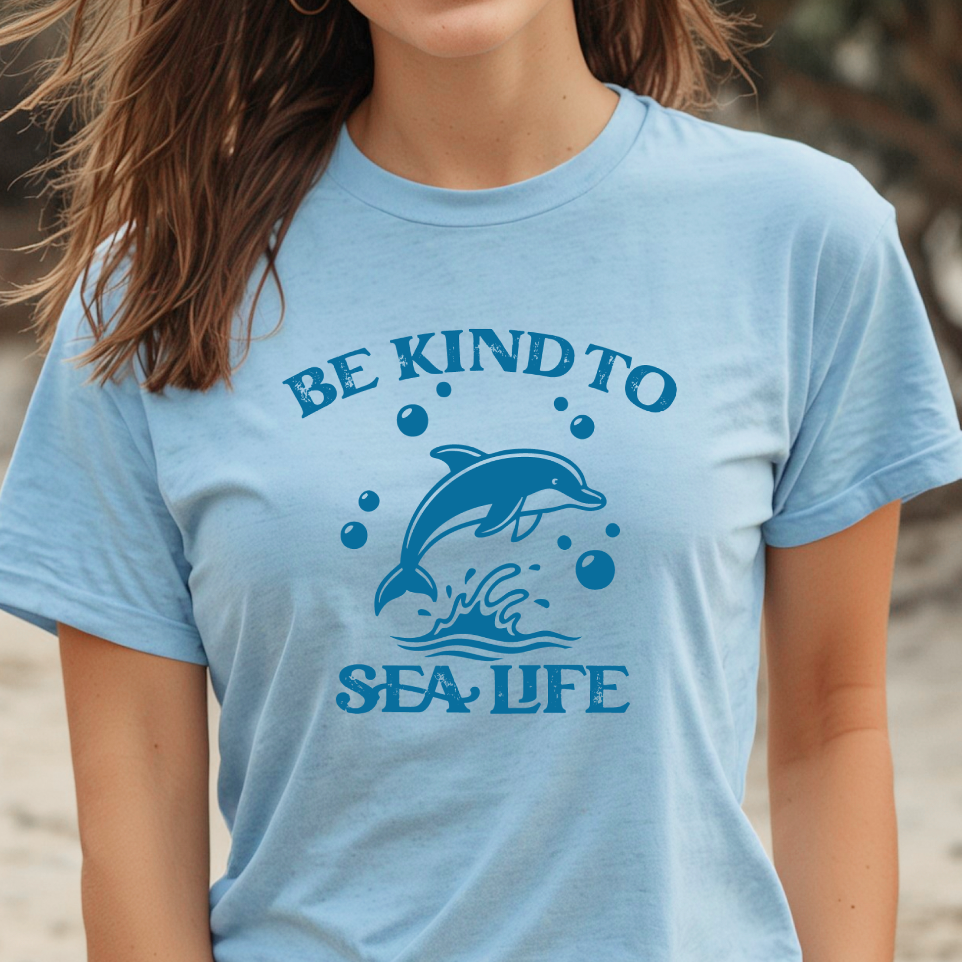 Person wearing a light blue t-shirt with a dolphin graphic and 'Be Kind to Sea Life' text on a beach.