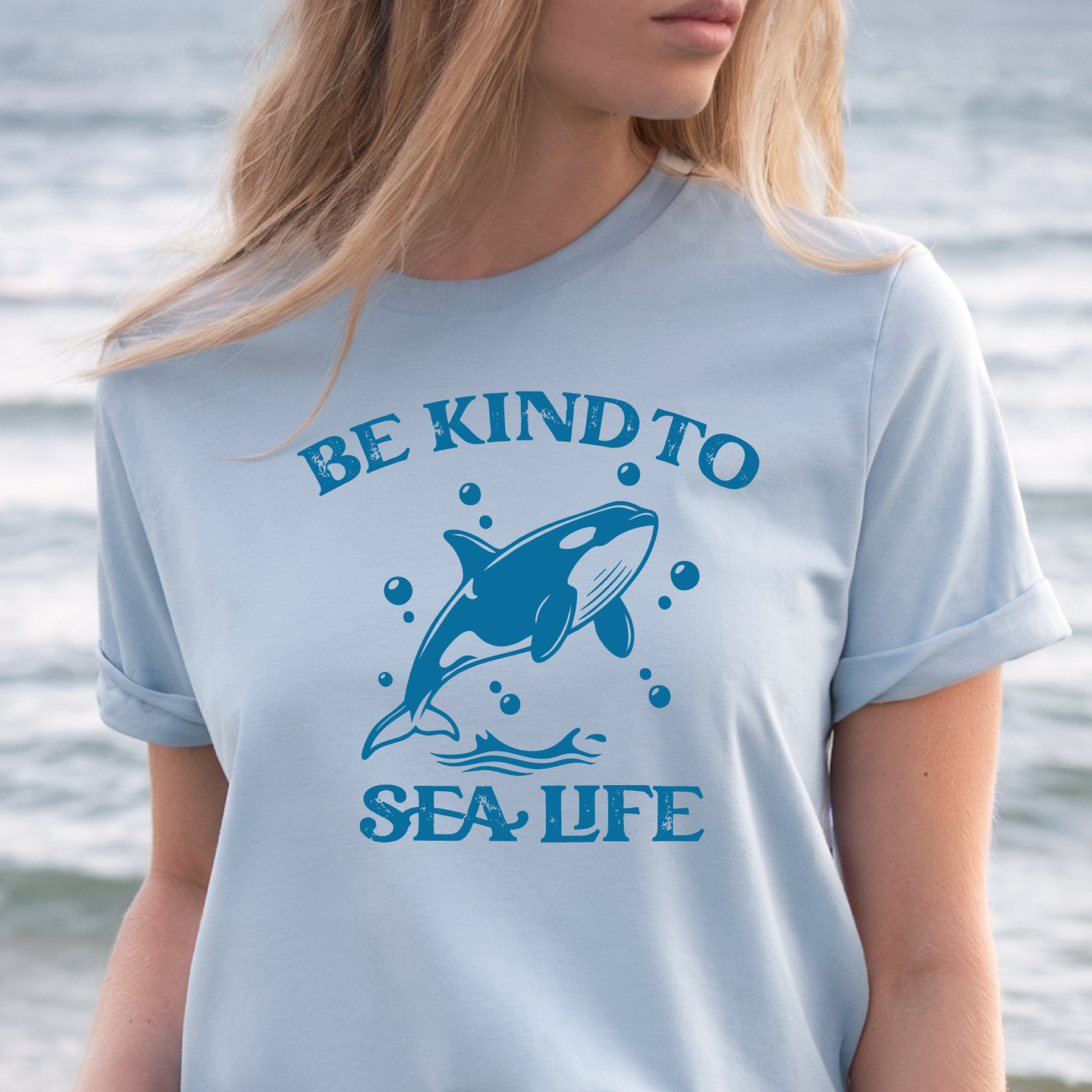 Person wearing a light blue t-shirt with a whale graphic and 'Be Kind to Sea Life' text by the water.