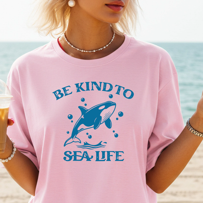 Person wearing a pink t-shirt with a whale graphic and 'Be Kind to Sea Life' text on a beach.