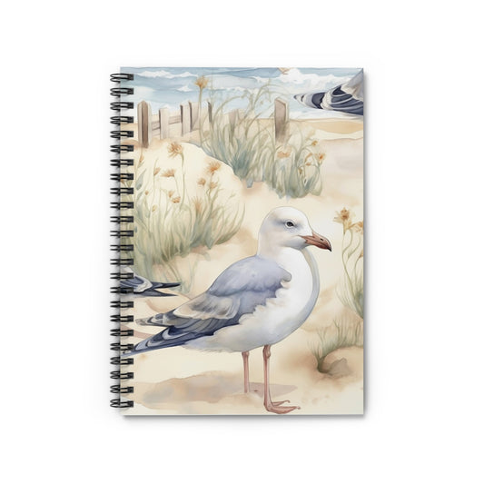 The Coastal Seagull Spiral Notebook features a cover with a seagull on a sandy beach with grass and a fence, under a partly cloudy sky, creating serene coastal charm perfect for beach lovers.
