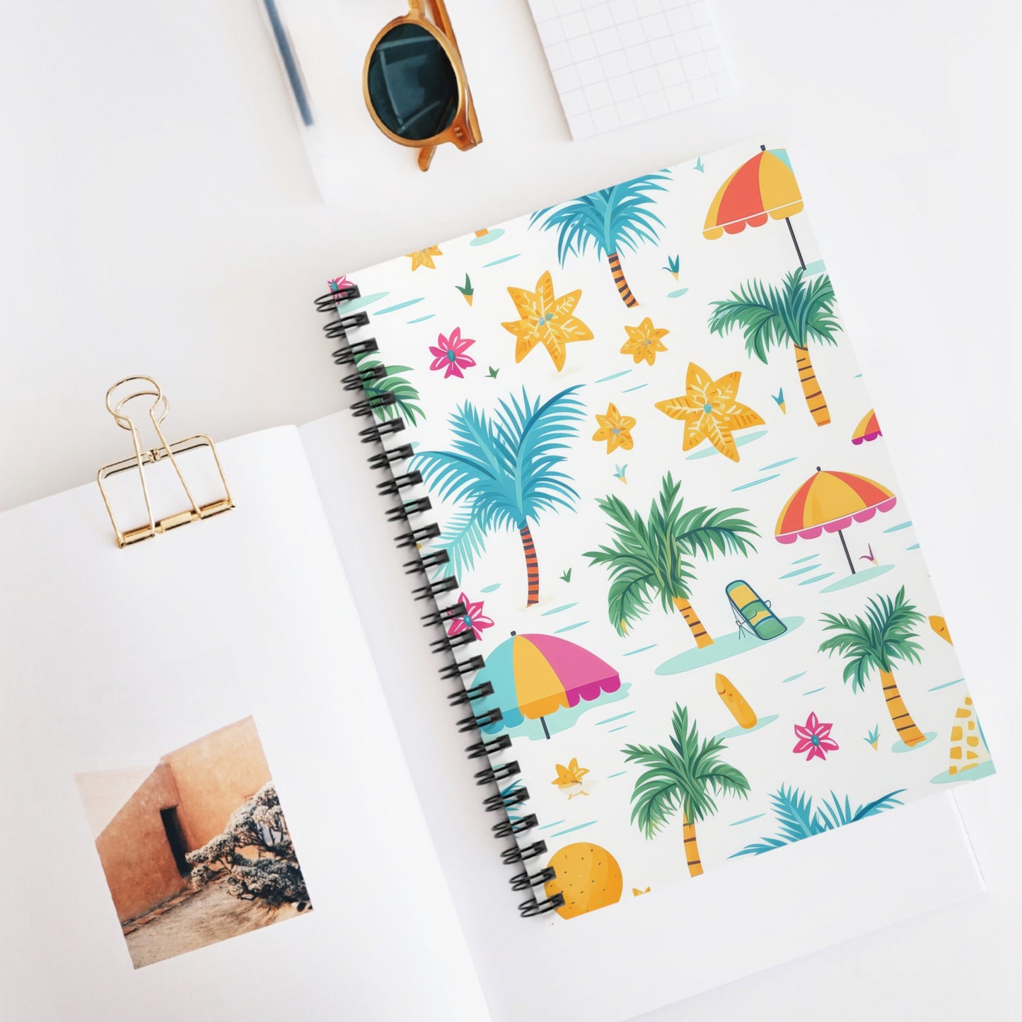 The Tropical Vibes Spiral Notebook, decorated with beach-themed graphics of palm trees, umbrellas, and flowers, rests on a white desk next to sunglasses, a binder clip, a notepad, and an open book with a photo—ideal for motivational journaling in sunny weather.