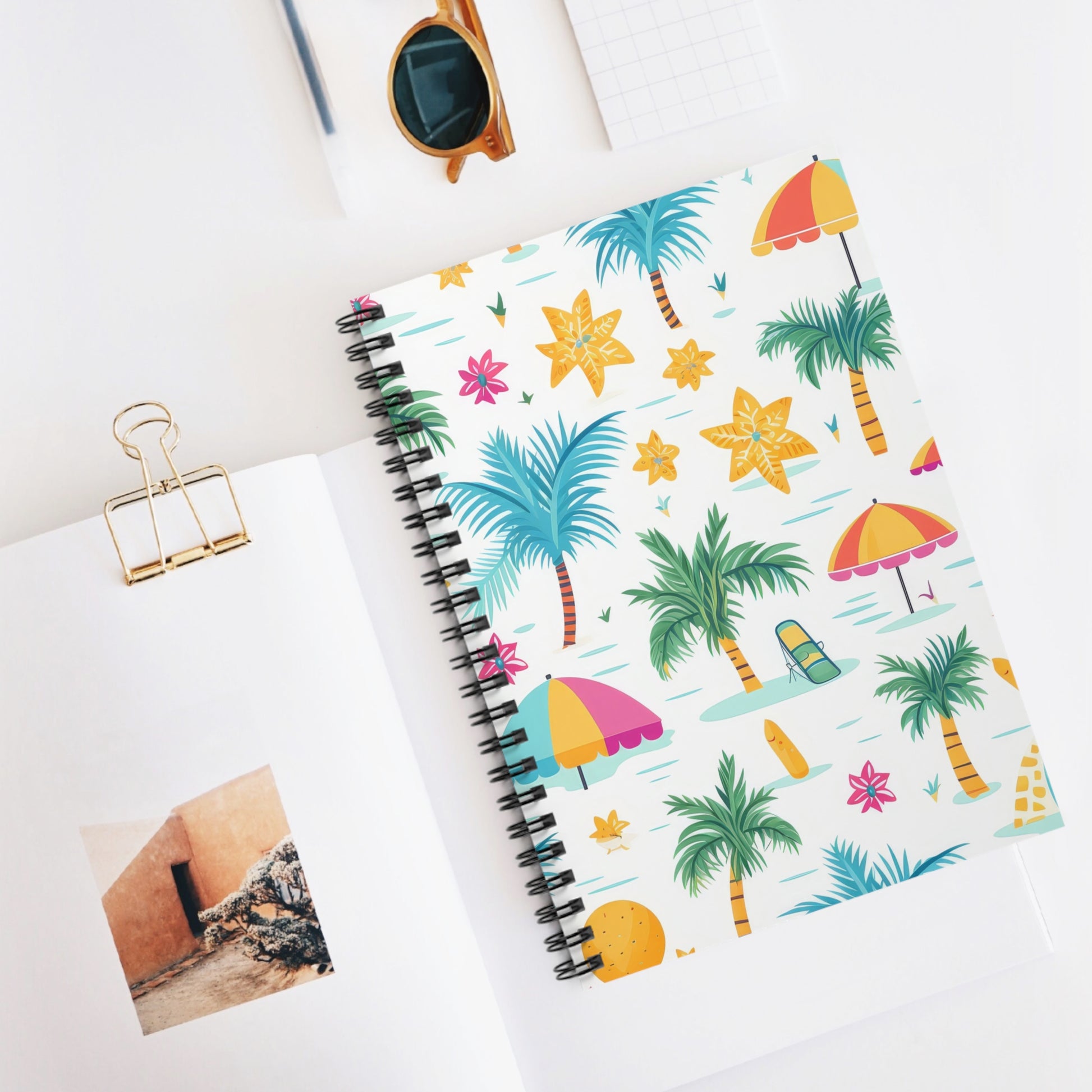 The Tropical Vibes Spiral Notebook, decorated with beach-themed graphics of palm trees, umbrellas, and flowers, rests on a white desk next to sunglasses, a binder clip, a notepad, and an open book with a photo—ideal for motivational journaling in sunny weather.