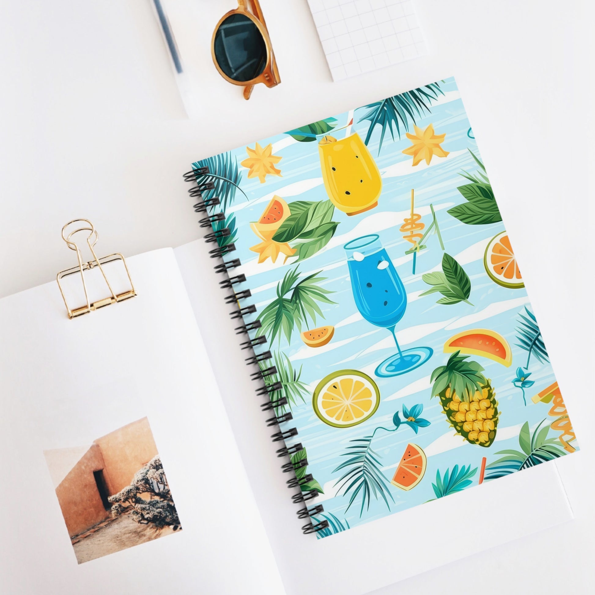 The Tropical Vibes Spiral Notebook, ideal for summer inspiration, sits on a white surface. Nearby, an open book with architecture imagery, brown sunglasses, and a small grid notebook ignite creative journaling.