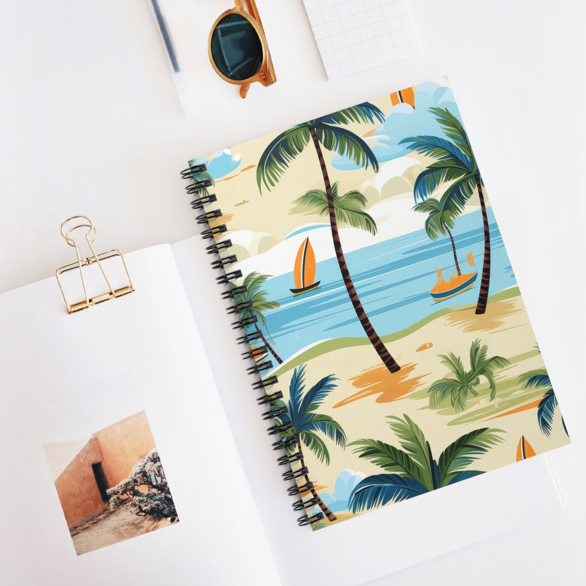 The Tropical Paradise Spiral Notebook - Ruled Line, featuring palm trees and boats on its cover, is open on a white surface. Nearby rest sunglasses, a small grid notebook, and a binder clip. A partial art print of a wall with steps is slightly visible.