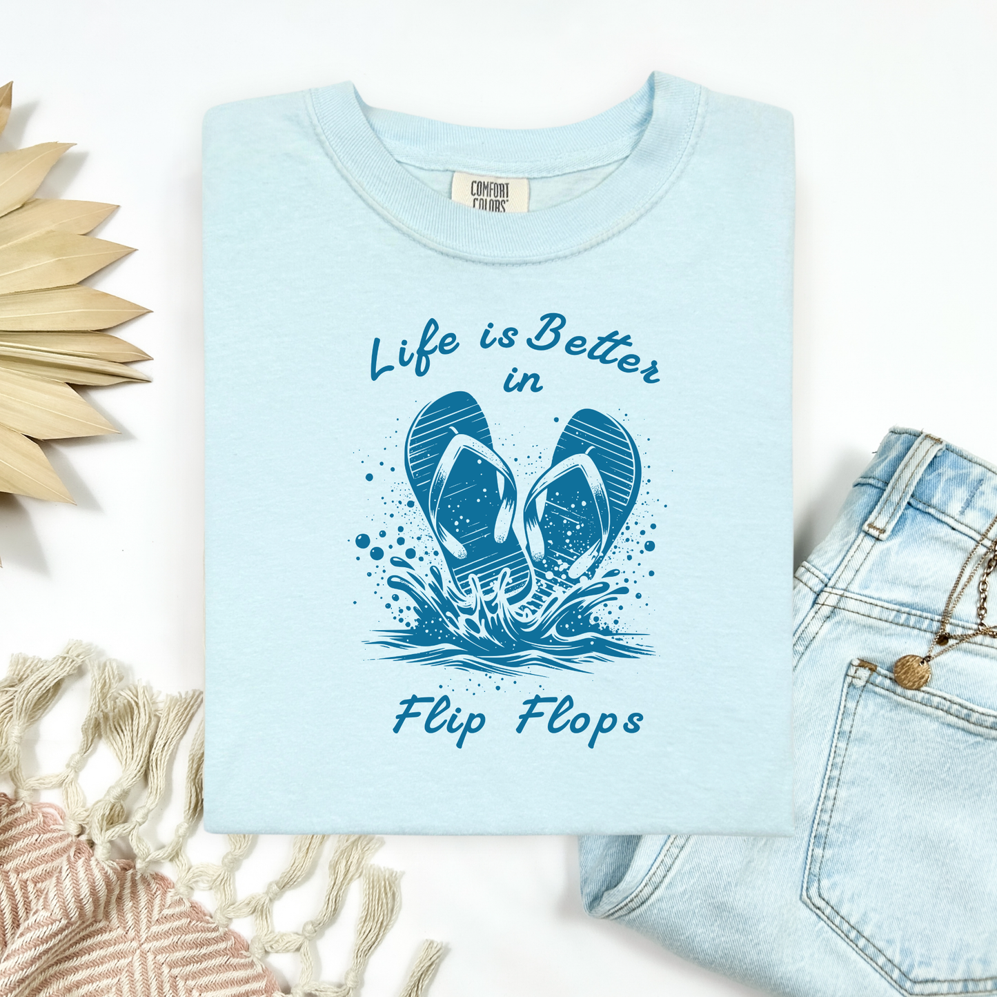 Comfort Colors 1717 "Life is Better in Flip Flops" t shirt with a pair of flip flops splashing in water one color graphic. Picture on a flay lay mockup with pair of blue jeans and blanket and a wicker sun shaped accessory.