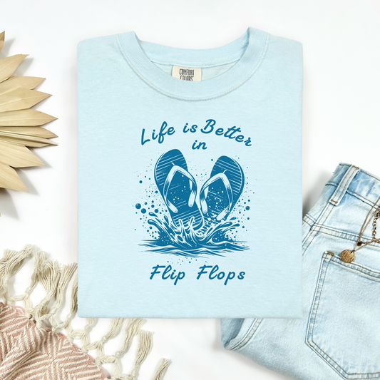 Comfort Colors 1717 "Life is Better in Flip Flops" t shirt with a pair of flip flops splashing in water one color graphic. Picture on a flay lay mockup with pair of blue jeans and blanket and a wicker sun shaped accessory.