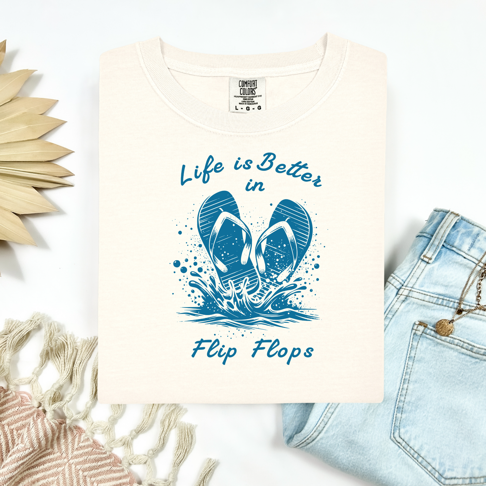 Comfort Colors 1717 "Life is Better in Flip Flops" t shirt with a pair of flip flops splashing in water one color graphic. Picture on a flay lay mockup with pair of blue jeans and blanket and a wicker sun shaped accessory.