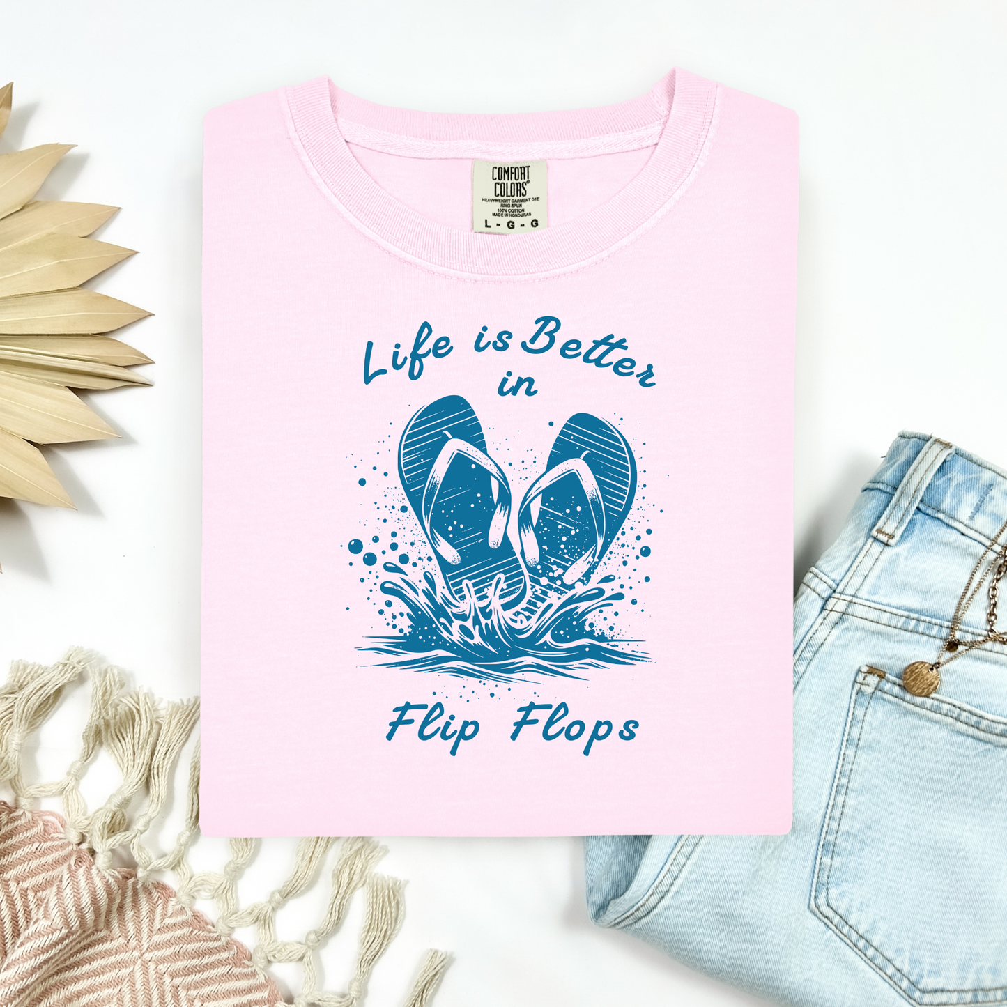 Comfort Colors 1717 "Life is Better in Flip Flops" t shirt with a pair of flip flops splashing in water one color graphic. Picture on a flay lay mockup with pair of blue jeans and blanket and a wicker sun shaped accessory.