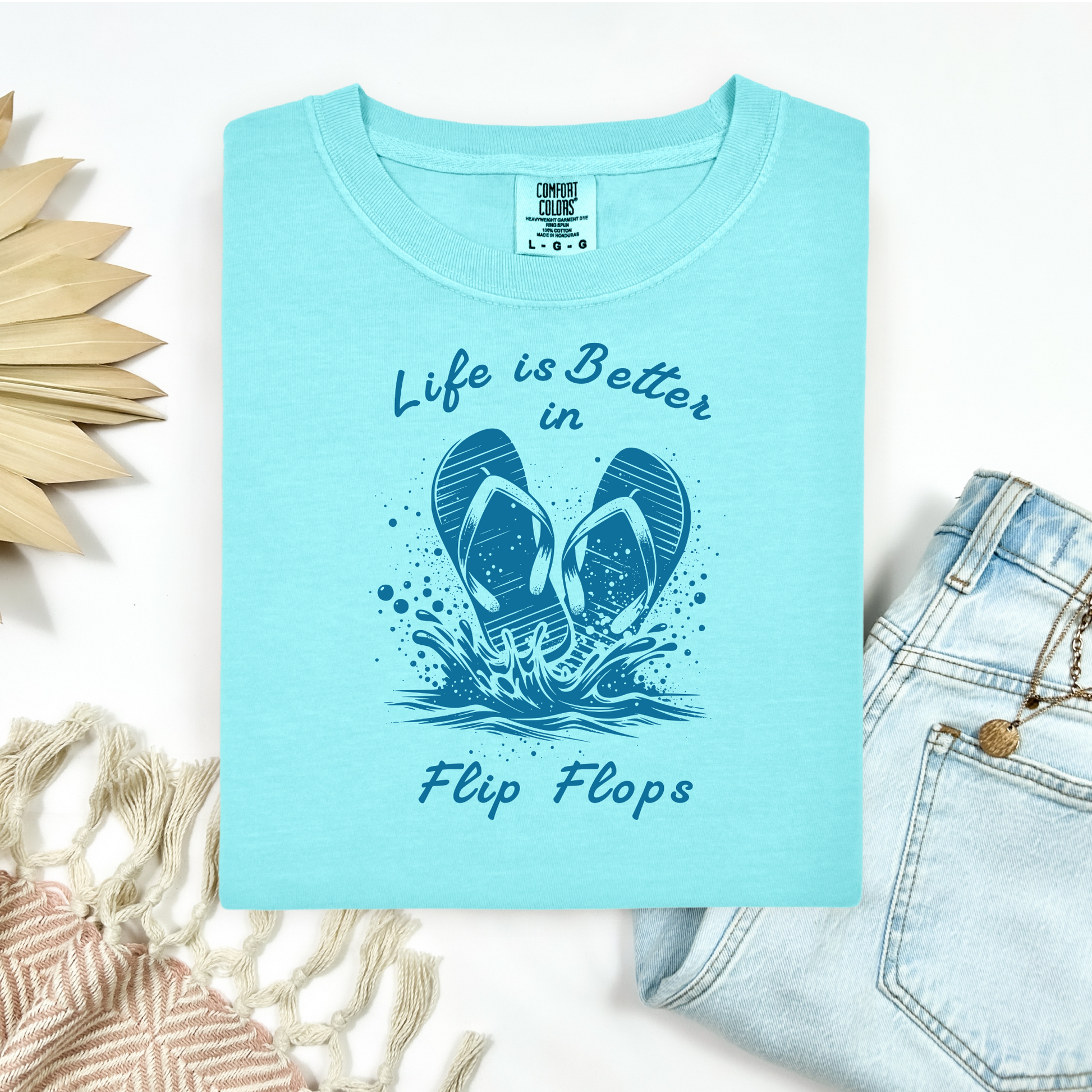 Comfort Colors 1717 "Life is Better in Flip Flops" t shirt with a pair of flip flops splashing in water one color graphic. Picture on a flay lay mockup with pair of blue jeans and blanket and a wicker sun shaped accessory.