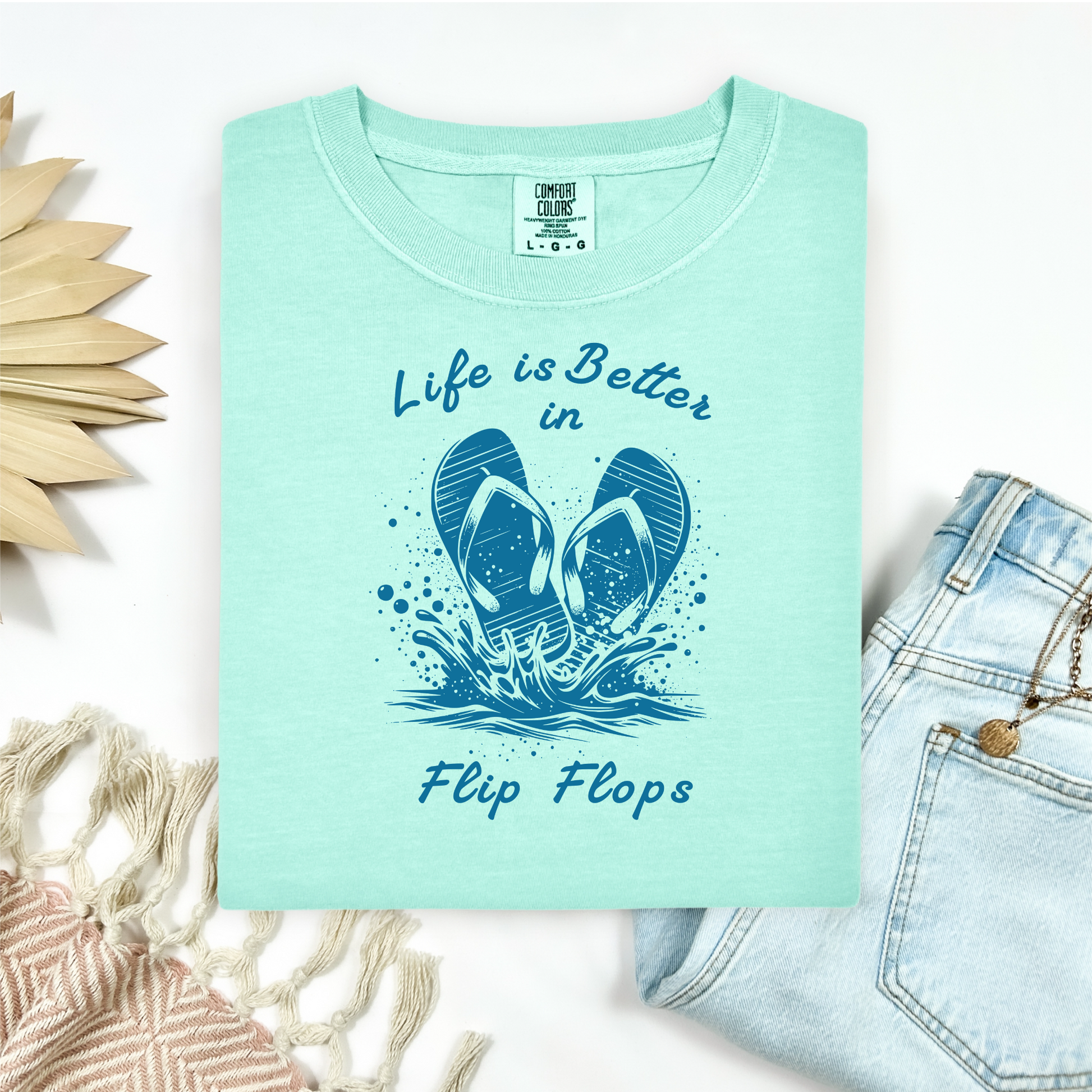 Comfort Colors 1717 "Life is Better in Flip Flops" t shirt with a pair of flip flops splashing in water one color graphic. Picture on a flay lay mockup with pair of blue jeans and blanket and a wicker sun shaped accessory.